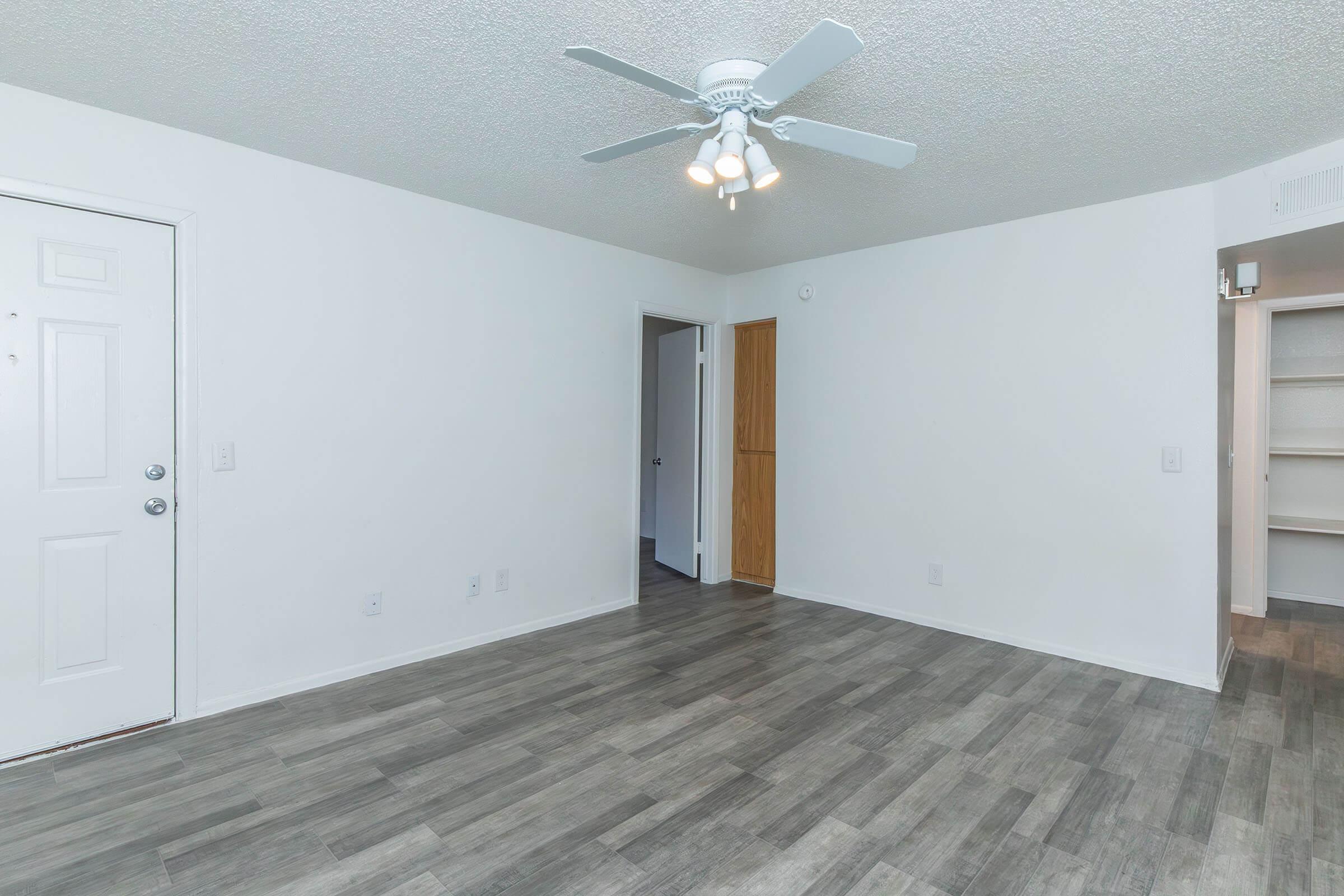 Empty room with light-colored walls and a textured ceiling. A ceiling fan with five lights is visible. An entry door is on the left, and a doorway leading to another room is in the center. The flooring is dark gray, and there's a small closet in the background. Natural light may come from a window not shown in the image.