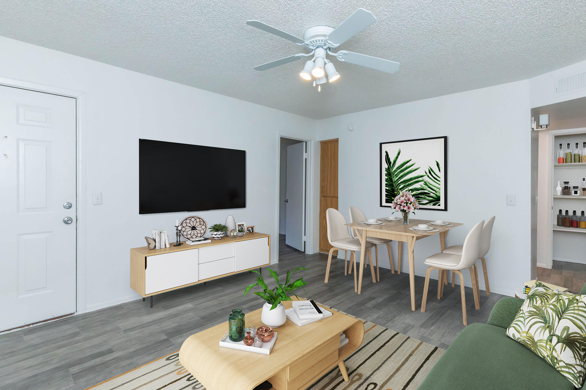 A modern living room featuring light-colored walls, a large flat-screen TV mounted on the wall, a wooden coffee table with decorative items, and a dining area with a simple table and chairs. There is a plant in a pot and artwork depicting leaves on the wall. The flooring is gray tiles, and there is a ceiling fan.