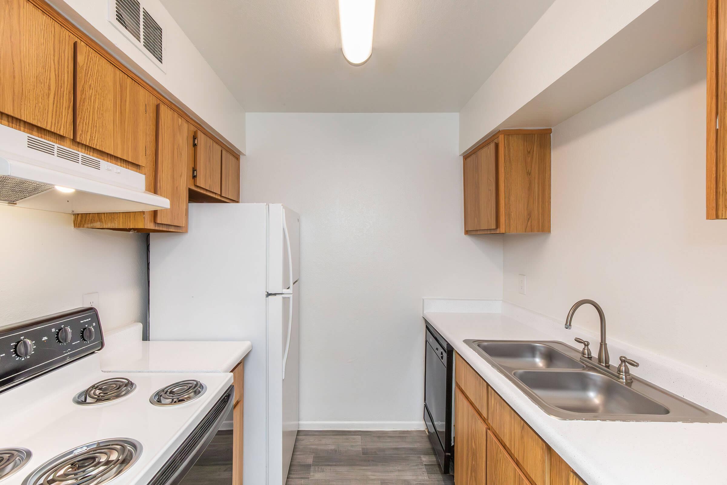 A modern kitchen featuring light wood cabinets, a white refrigerator, a silver stove, and a double sink. The countertops are white, and the flooring is a light gray. A ceiling light provides illumination, and the walls are painted a neutral color. The overall design is clean and functional.