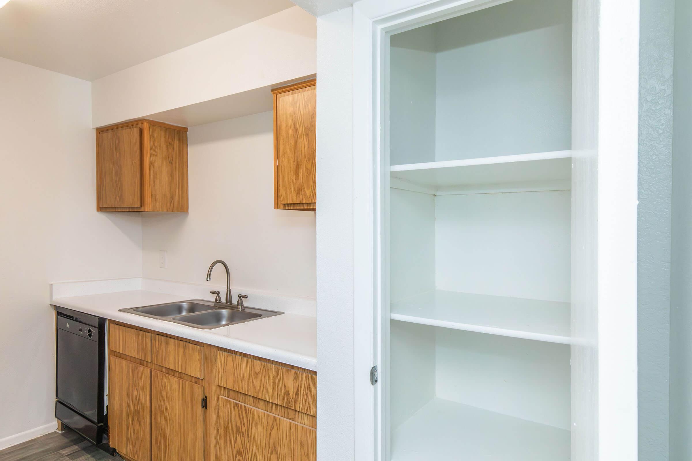 A kitchen area featuring stainless steel appliances, including a dishwasher, and wooden cabinets. On the right, there is a white pantry or storage closet with empty shelves. The walls are painted white, and the floor has a simple tile pattern.