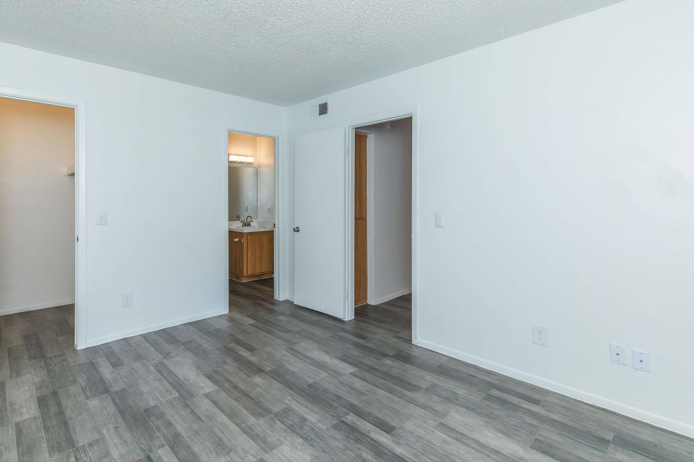 Interior view of a room with light gray wood-like flooring and white walls. Two doorways are visible, one leading to a bathroom with a vanity, and the other leading to a closet. The overall space is bright and uncluttered, suggesting a modern design.