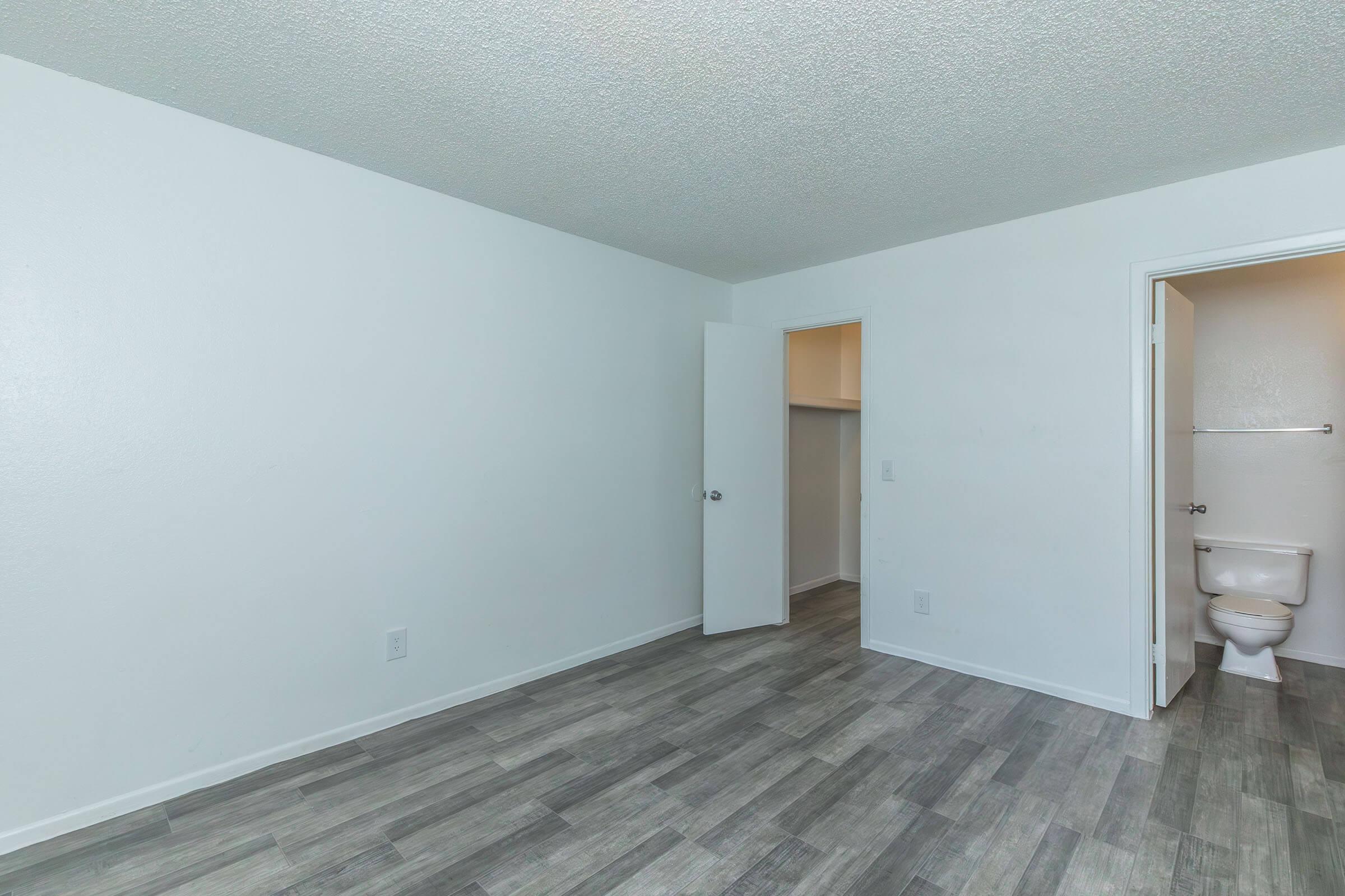 A spacious, empty room with light gray wood-style flooring. The walls are painted white, and there are two doors: one leading to a small bathroom and another leading to another area. The room has a simple and clean appearance, suitable for various uses.
