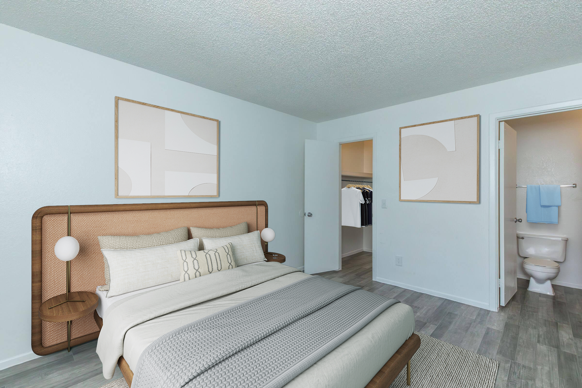 A modern bedroom featuring a double bed with a light gray blanket, two decorative pillows, and a rattan headboard. The walls are painted light blue, adorned with two abstract art pieces. There’s a closet in the background and an adjoining bathroom with a toilet visible. The flooring is a light wood texture.