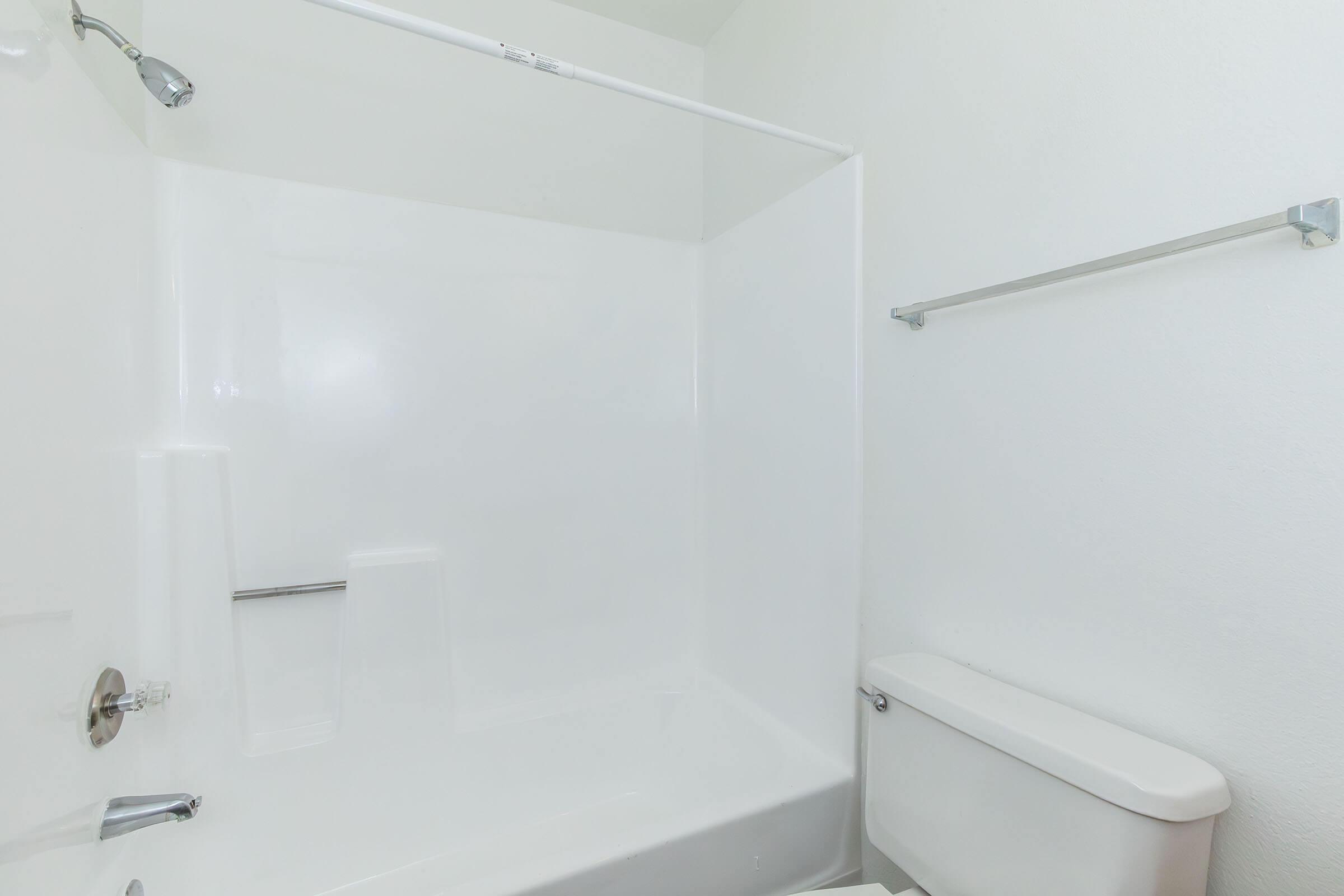 A clean and empty bathroom featuring a white shower stall with a showerhead, a bathtub with a curved shower rod, a white toilet, and a towel rack on the wall. The walls and fixtures are bright and minimalist, creating a spacious and fresh appearance.