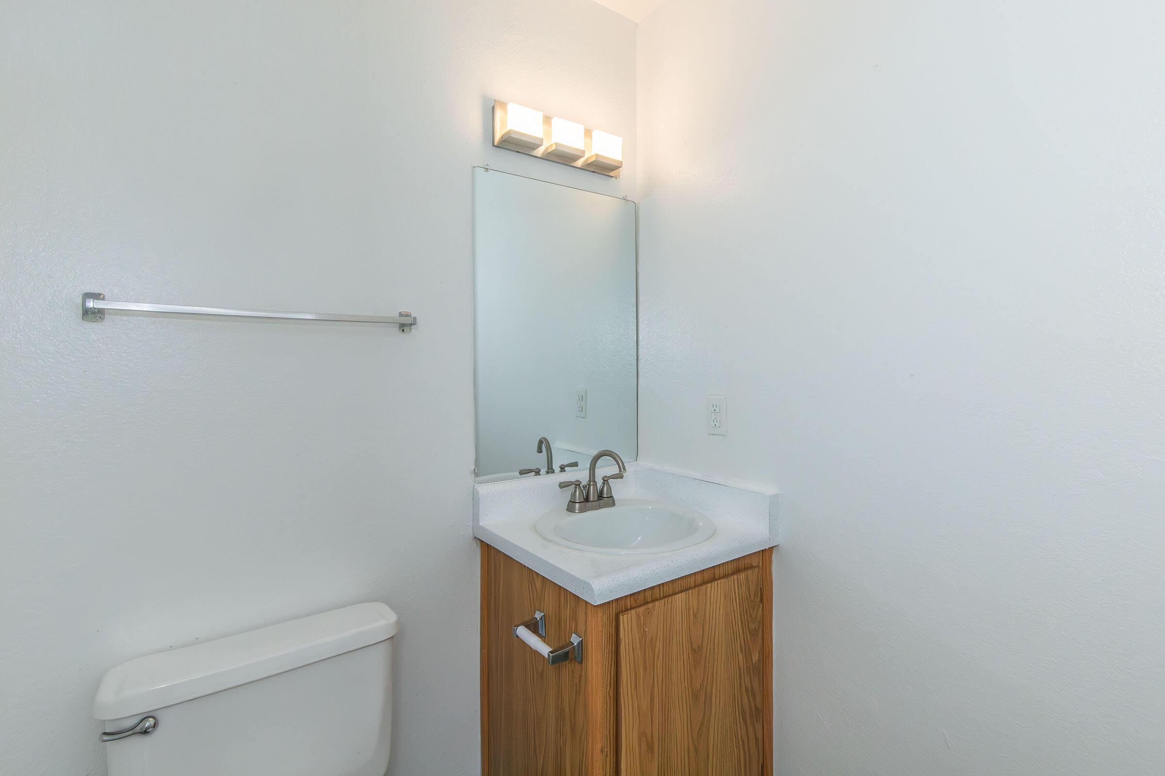 A clean and simple bathroom featuring a white sink with a wooden cabinet below, a rectangular mirror above, a wall-mounted light fixture with multiple bulbs, and a standard toilet. The walls are painted white, creating a bright and open feel.