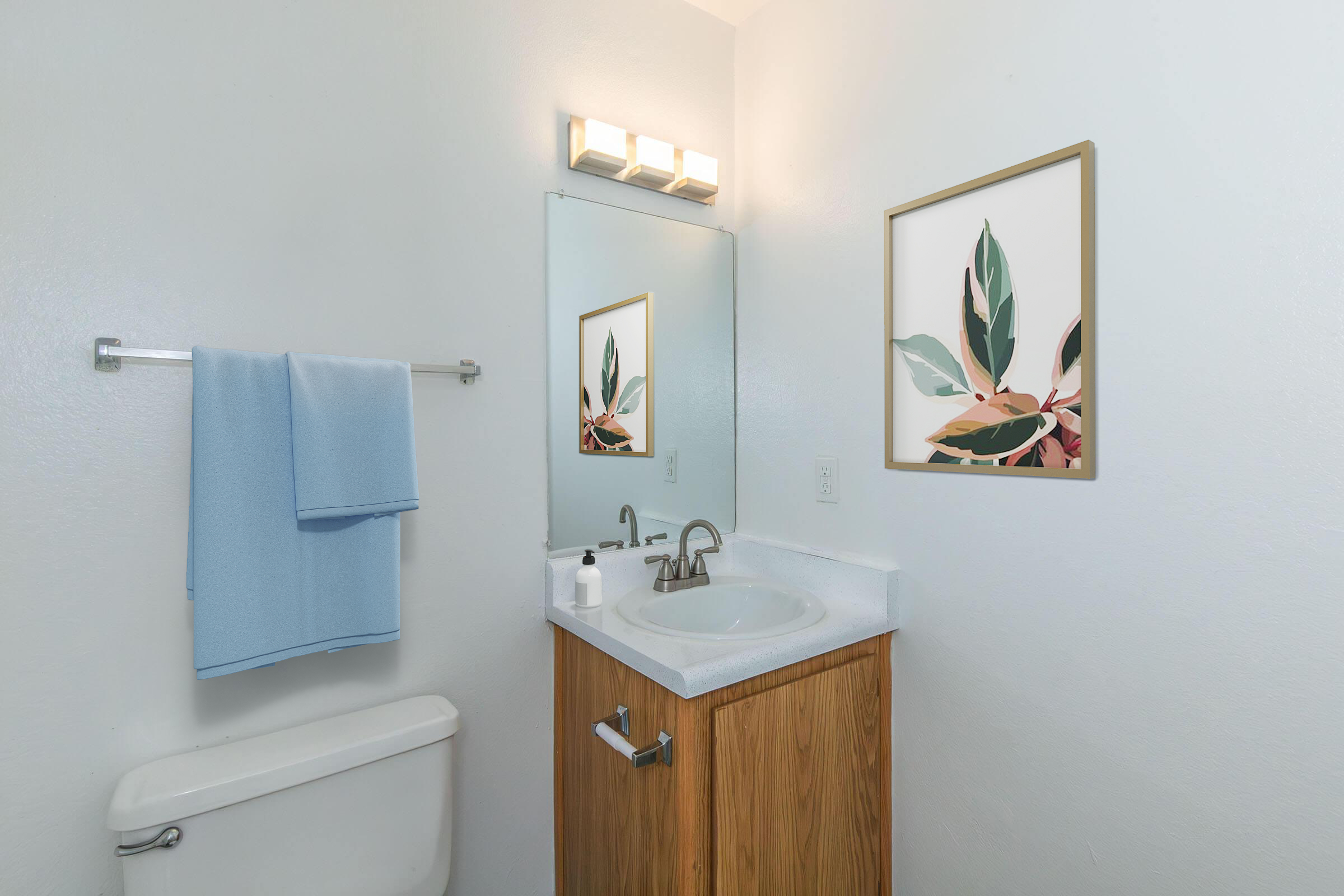 A well-lit bathroom featuring a white sink with a silver faucet, a wooden cabinet below, and a towel rack with two light blue towels hanging. A large mirror reflects the space, and a decorative framed artwork of leaves is mounted on the wall. A white toilet is visible on the left.