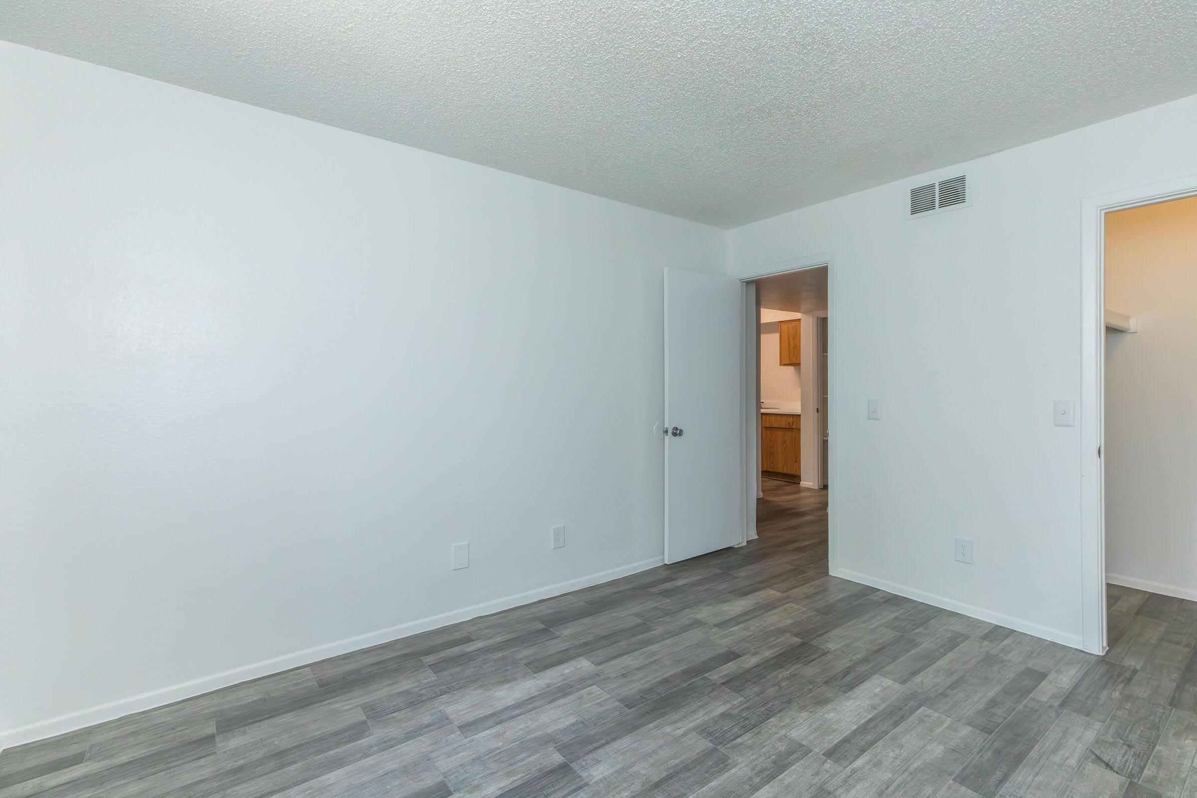 Empty room with light gray walls and laminate flooring. A door is slightly ajar on the left leading to a kitchen area. A closet door is visible on the right. The space is well-lit, creating a clean and open atmosphere, ready for furnishing.
