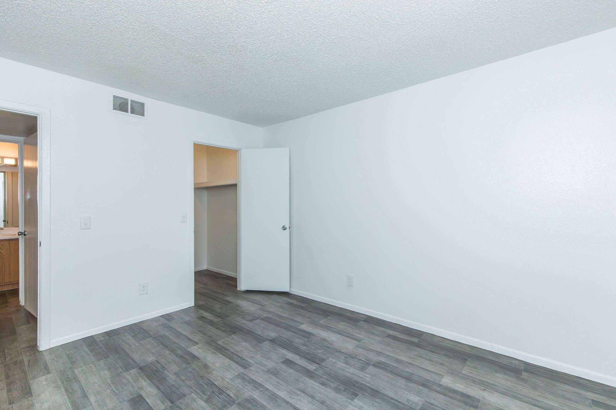 An empty room featuring a light gray tiled floor and white walls. On the left, a door leads to a bathroom, while an adjacent door is slightly ajar, revealing a small closet area. Natural light fills the space, creating a bright and open atmosphere.