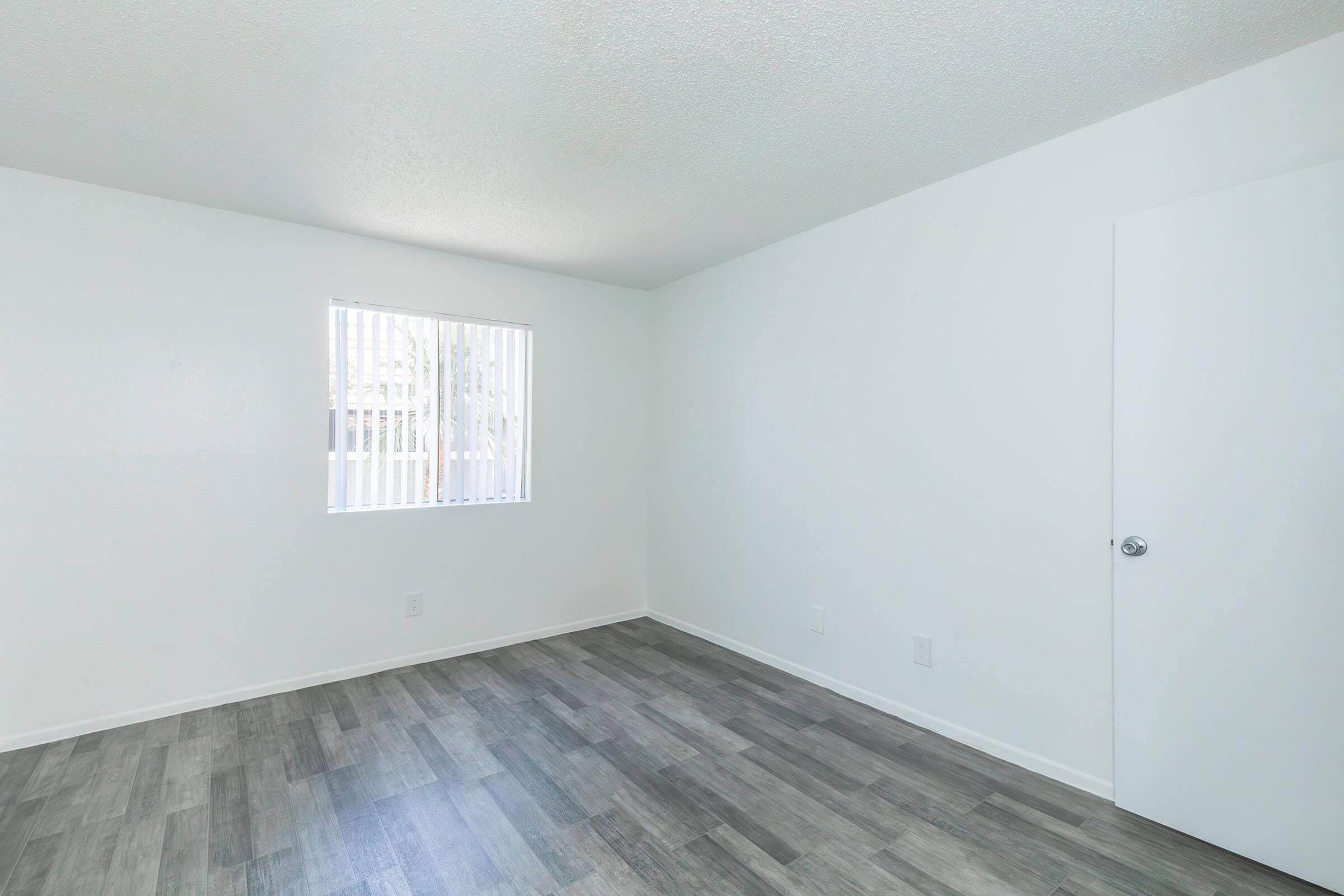 Empty room with white walls and wood-style flooring. A small window with vertical blinds allows natural light to enter. The room features a plain door and no furniture, creating a minimalistic and clean appearance. Ideal for various purposes, such as an office or guest room.