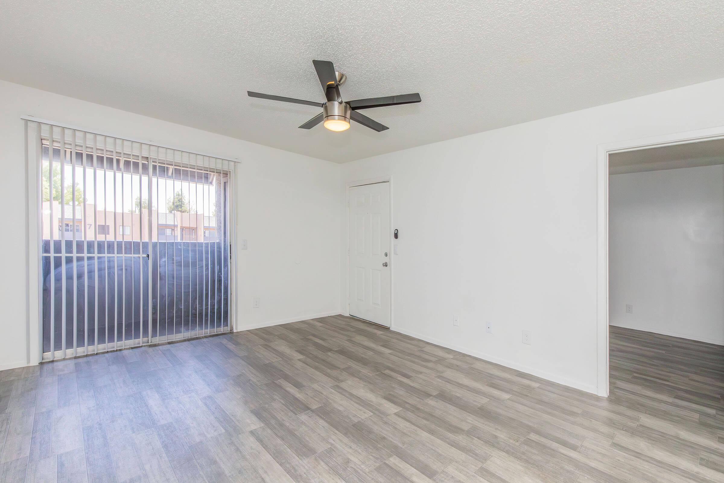A spacious, empty living room featuring light-colored walls, a ceiling fan, and large windows with vertical blinds. The floor is covered in light wood-like laminate, and a doorway leads to another room. Natural light fills the area, giving it a bright and airy feel.