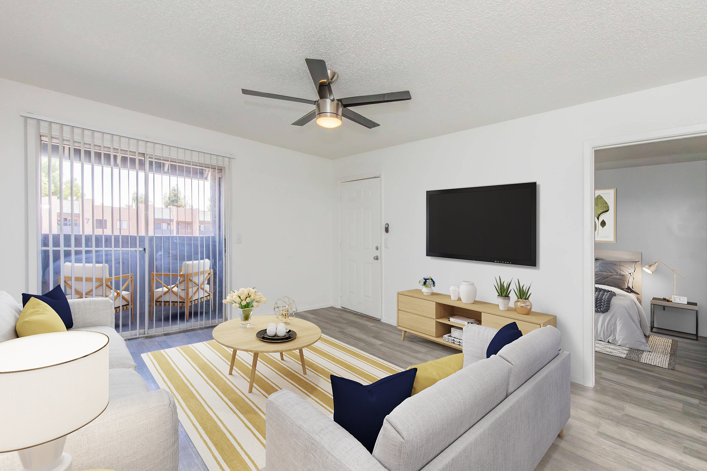 Bright and modern living room with a ceiling fan, featuring a light gray sofa and a round coffee table on a striped rug. A large wall-mounted TV and a wooden console are visible. Sliding glass doors lead to a balcony, with a small seating area outside. The room is decorated with plants and soft accent pillows.