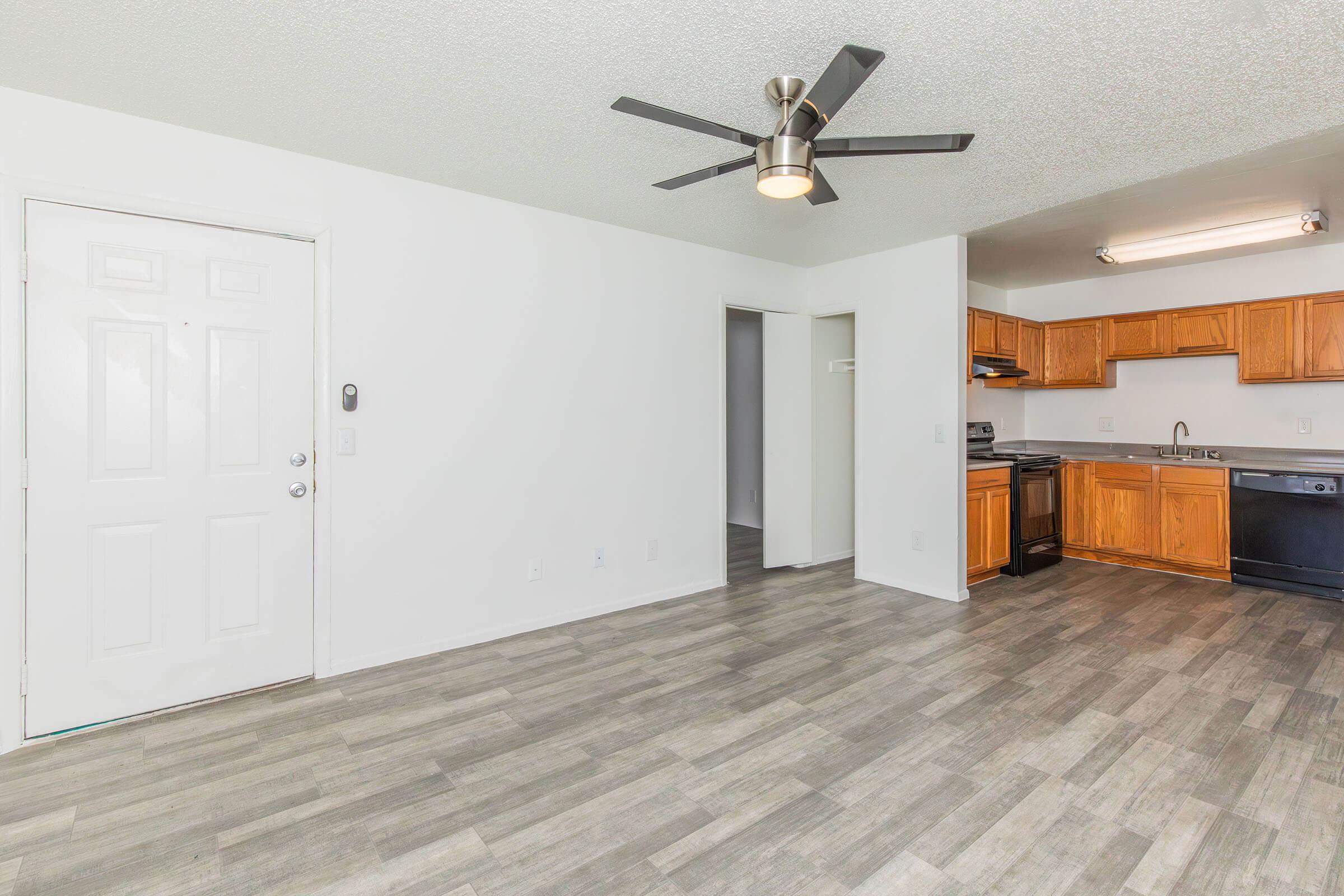Interior view of a modern apartment featuring a spacious living area with light-colored walls, a ceiling fan, and laminate flooring. The kitchen is partially visible, showcasing wooden cabinets, counter space, and appliances. There is a closed door leading to a room and another door leading outside.