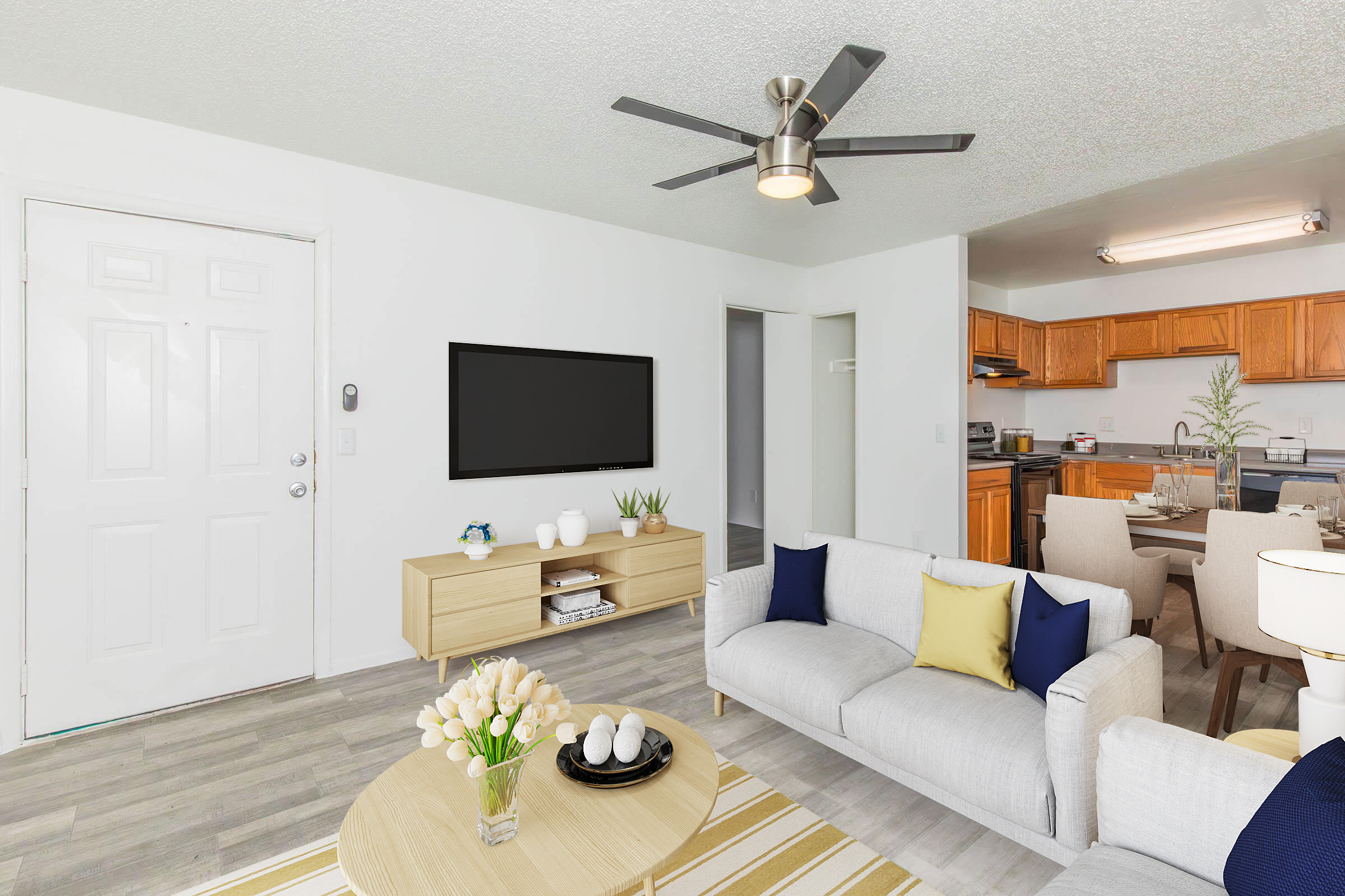 A bright and modern living room featuring a light gray sofa with blue and yellow accent pillows, a wooden coffee table with decorative items, and a flat-screen TV mounted on the wall. The room has a door leading outside and an open kitchen area visible in the background, with warm wood cabinetry and appliances.