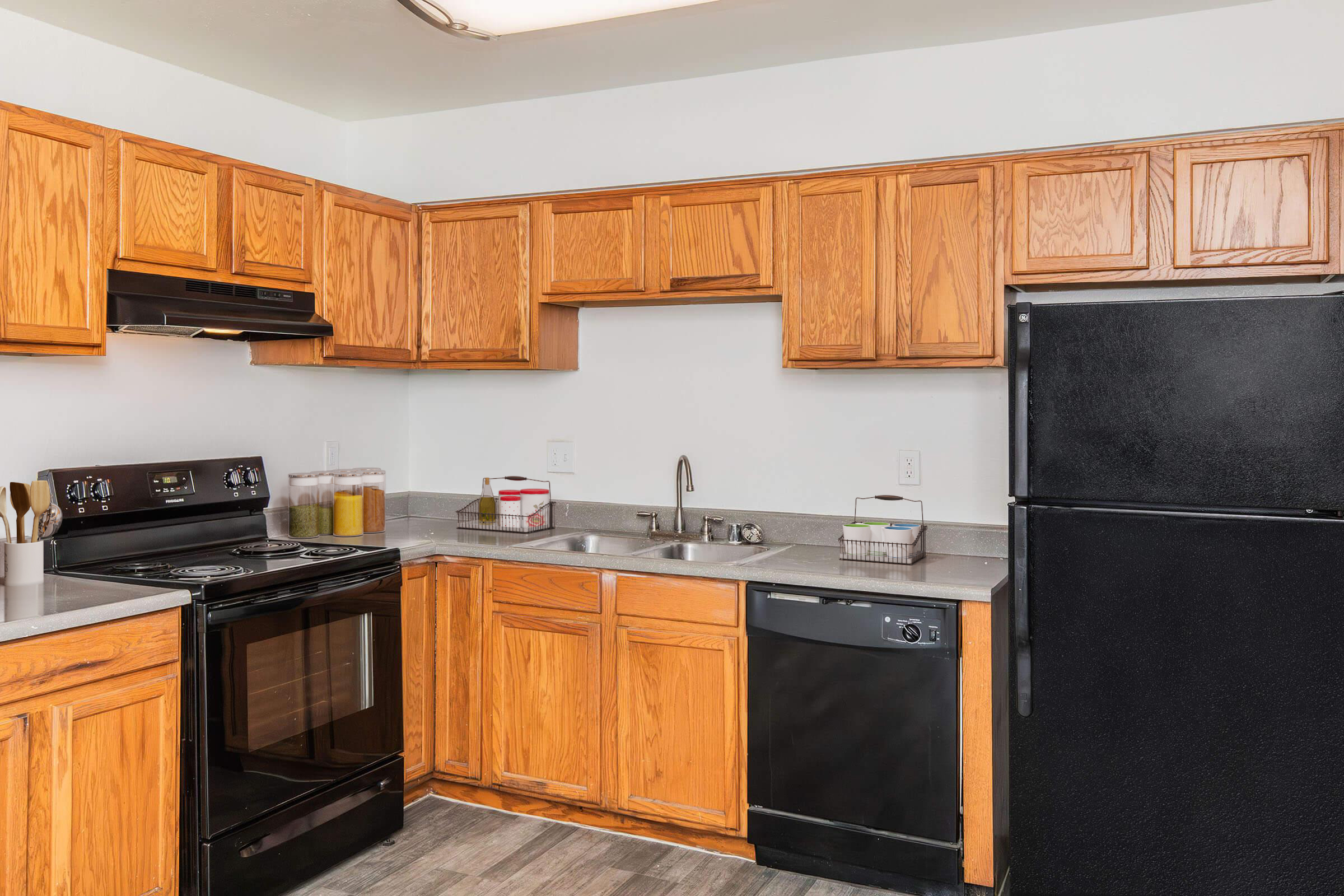 A modern kitchen featuring wooden cabinets, a black refrigerator, an oven, and a dishwasher. The countertop is equipped with a sink, and various utensils are organized on the counter. The flooring is grey, and there are containers of ingredients on the counter. The walls are plain and light-colored.