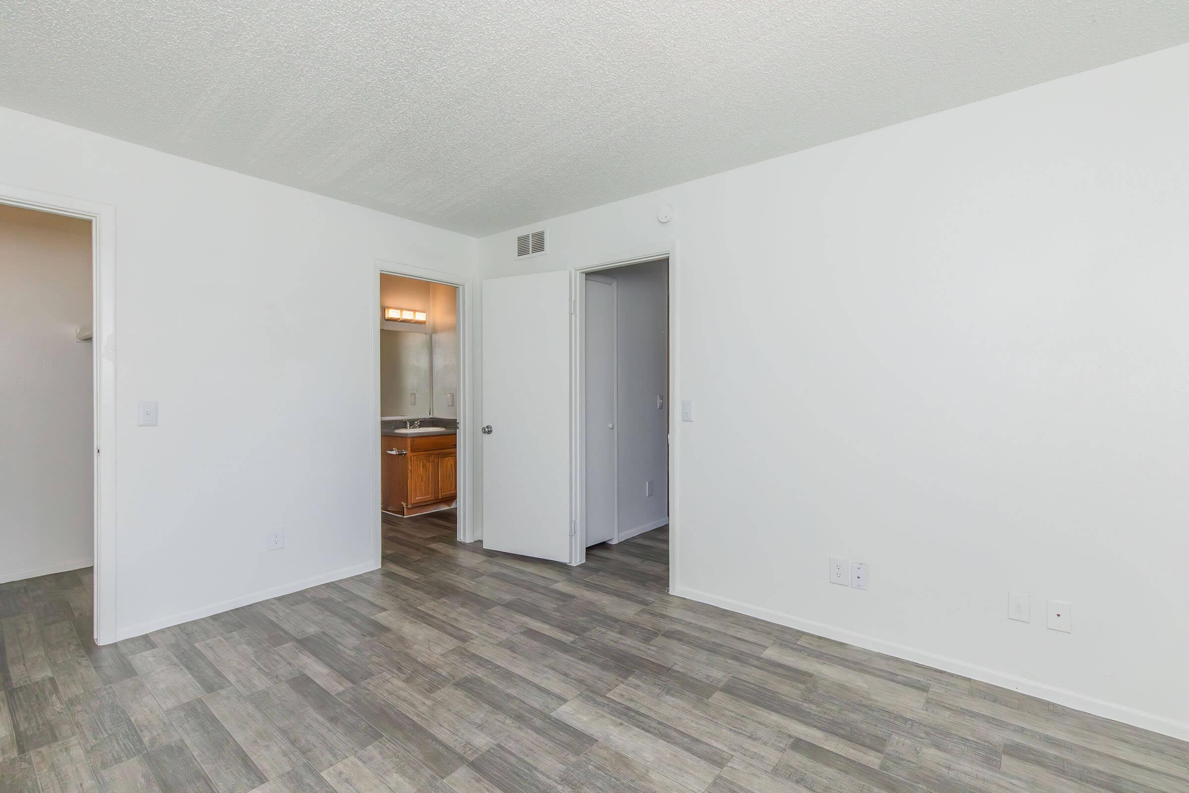 A bright, empty room with light-colored walls and a textured ceiling. The floor features grey laminate or tile. A doorway leads to a bathroom with wooden cabinetry visible. Another door provides access to an adjacent room. The overall atmosphere is clean and spacious.