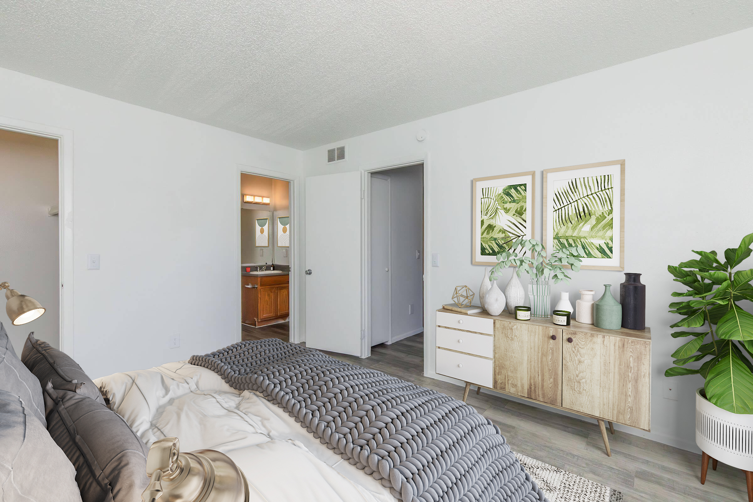 A cozy bedroom featuring a large bed with a grey blanket, a stylish wooden dresser with decorative vases and plants, and an adjoining doorway leading to a bathroom. The walls are painted white, and there are two framed botanical prints on the wall, adding a touch of greenery to the space.
