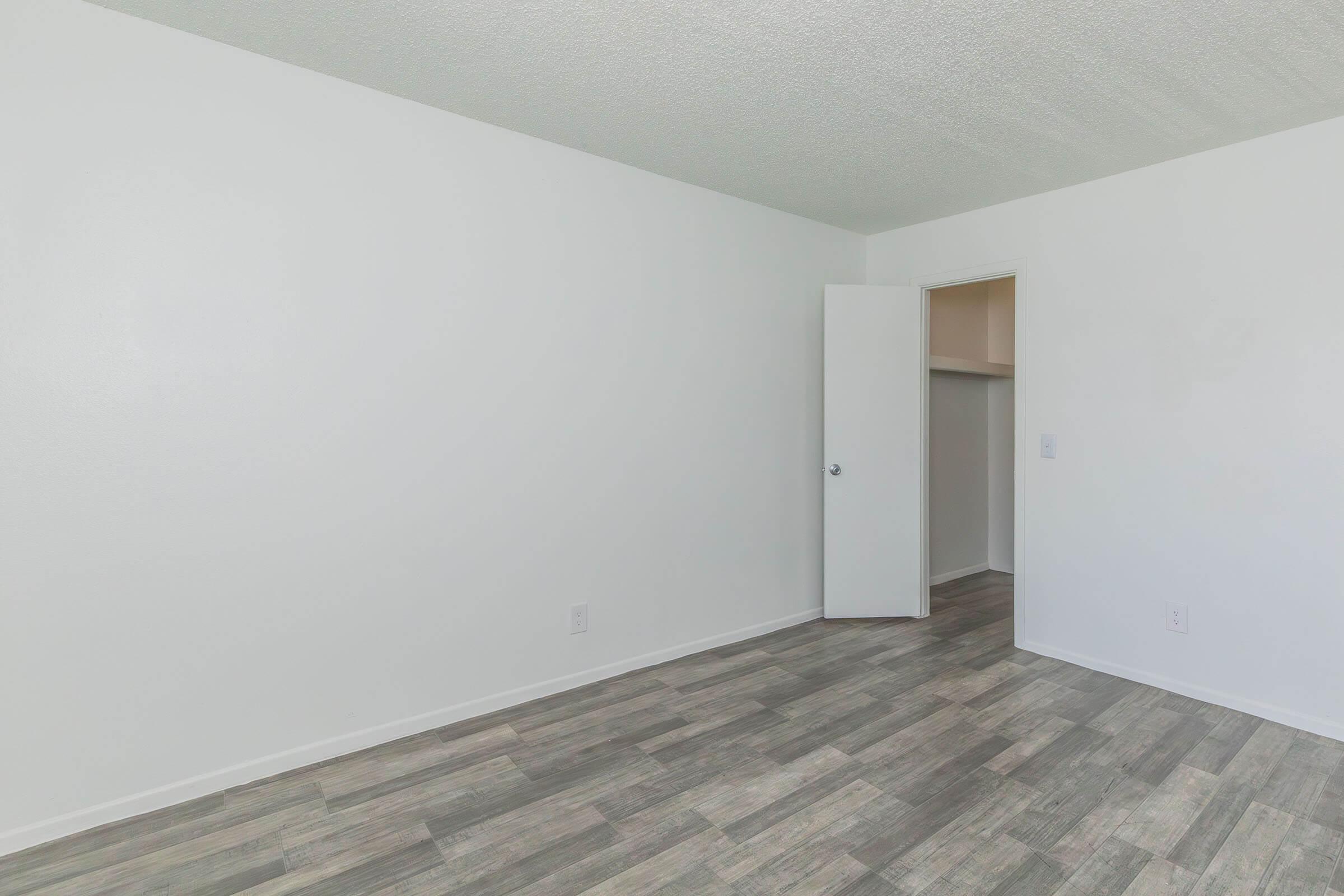 Empty room with white walls and a light-colored floor. One door is partially open, leading to a small closet area. The overall space appears bright and minimalistic, suitable for various uses or personalization.
