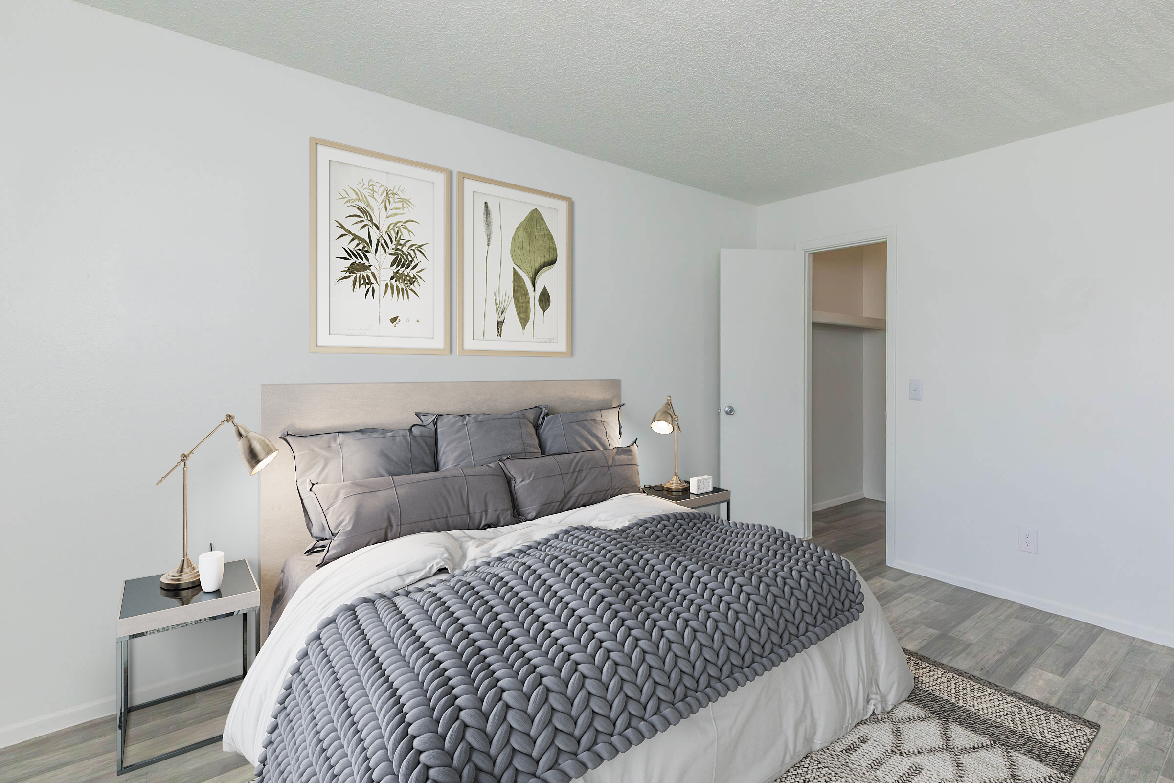 A modern bedroom featuring a queen-sized bed with gray bedding and decorative pillows. There are two framed botanical prints above the bed, matching bedside tables with lamps, and a light gray textured wall. A closet door is slightly ajar, revealing additional storage space. The flooring is light-colored wood.