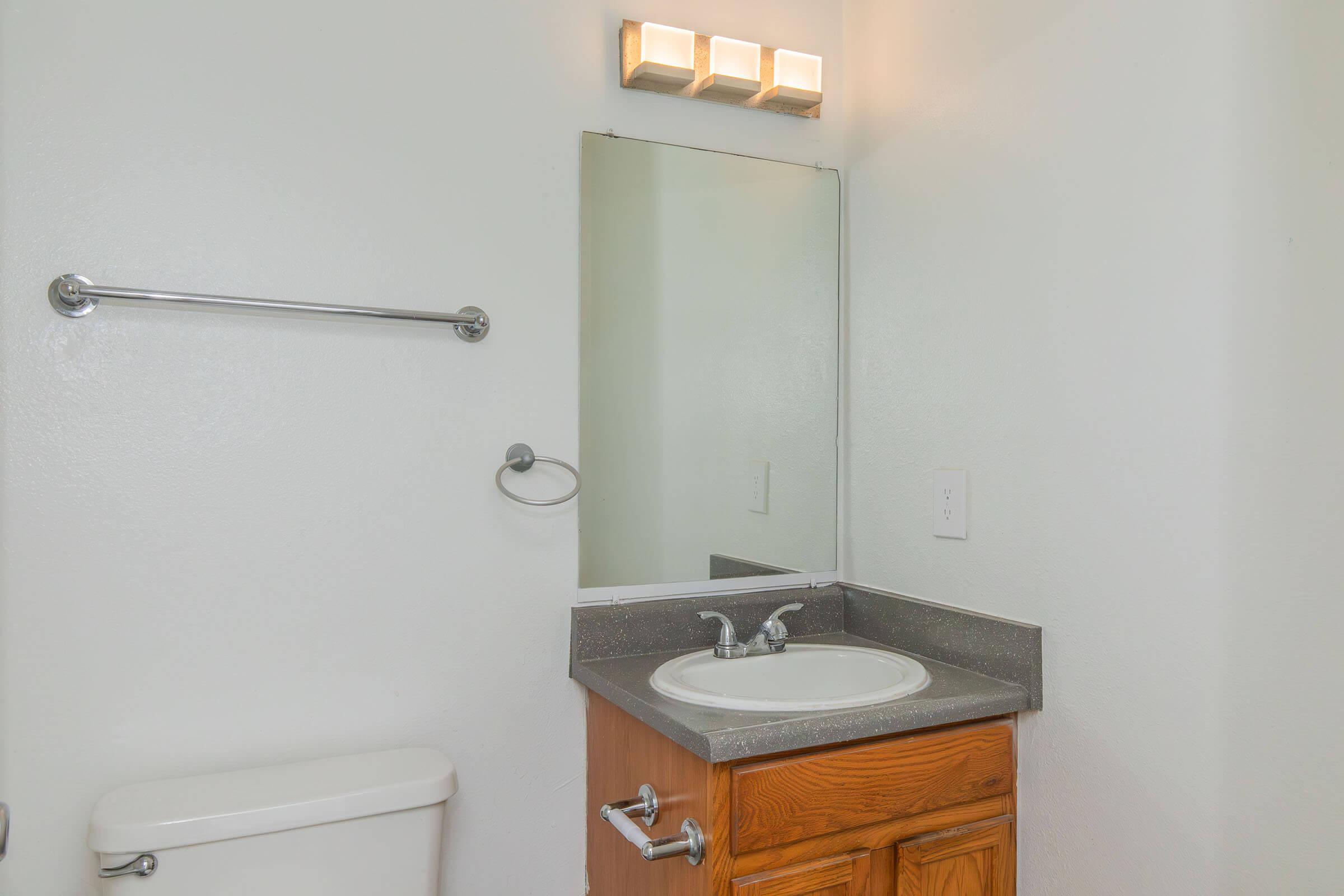 A clean bathroom with a small sink and wooden cabinet, a mirror above the sink, a towel rack on the wall, and a toilet nearby. The lighting is provided by a three-bulb fixture above the mirror, and the walls are painted white, contributing to a bright and spacious feel.
