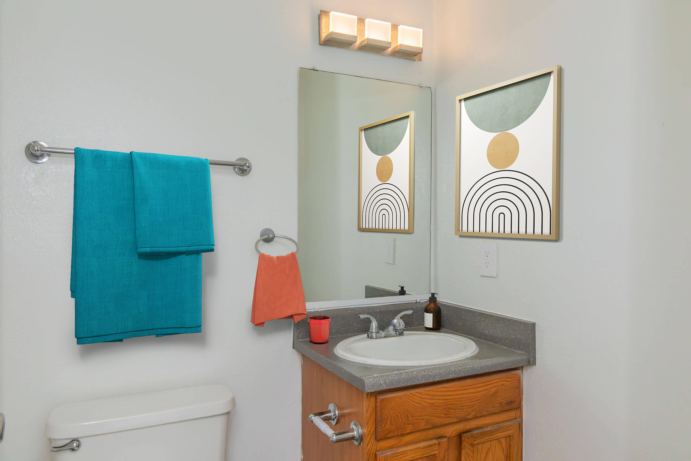 A bathroom featuring a wooden cabinet vanity with a sink, a mirror with a decorative frame, and two wall art pieces. There is a teal towel hanging on a silver rack and an orange hand towel on a hook. A white toilet is visible, and the walls are painted white, creating a bright and modern aesthetic.