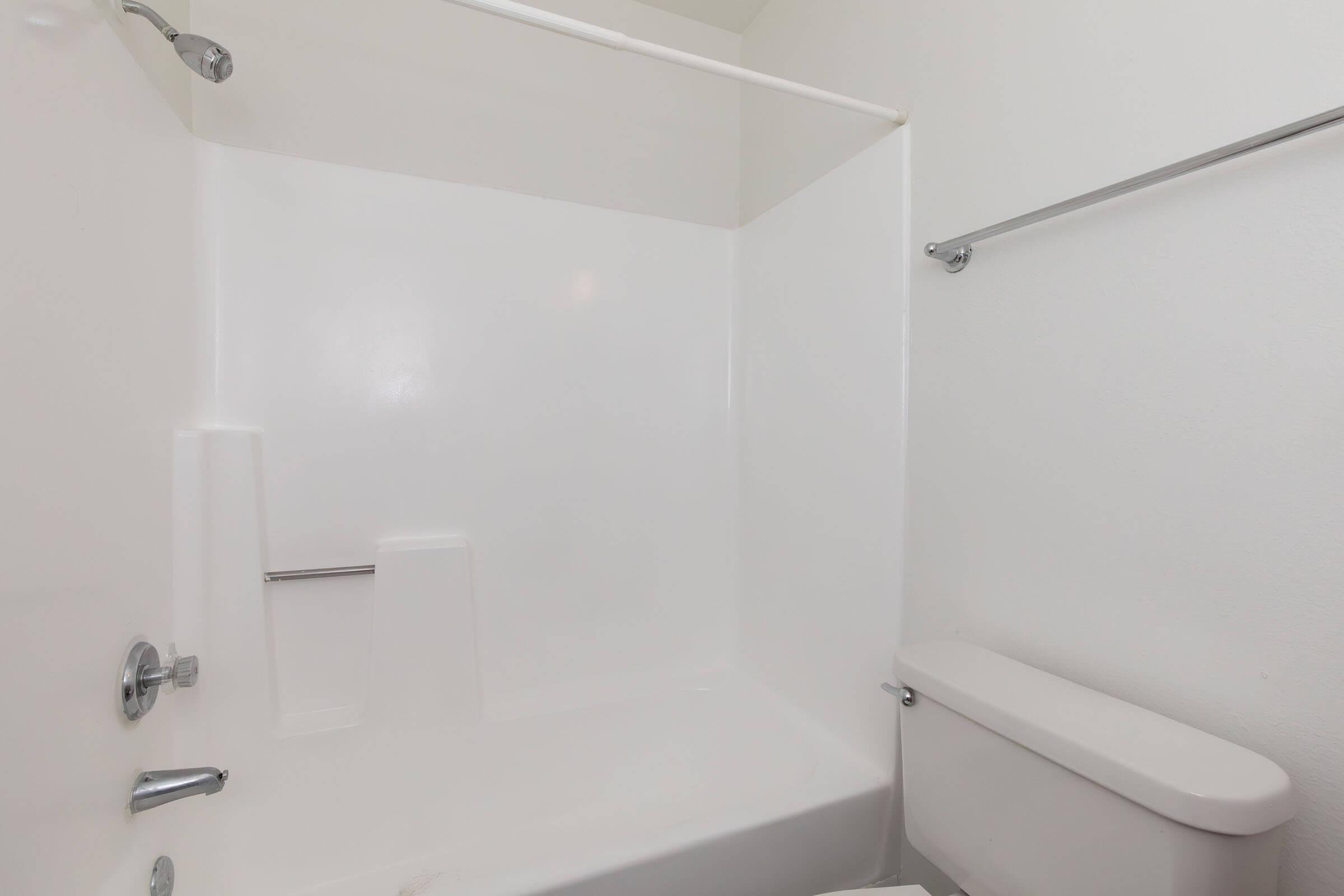 A clean and modern bathroom featuring a white bathtub with a shower curtain rod and a white toilet. The walls and fixtures are all white, creating a bright and spacious feel. The space is minimalistic with no additional decorations or items present.