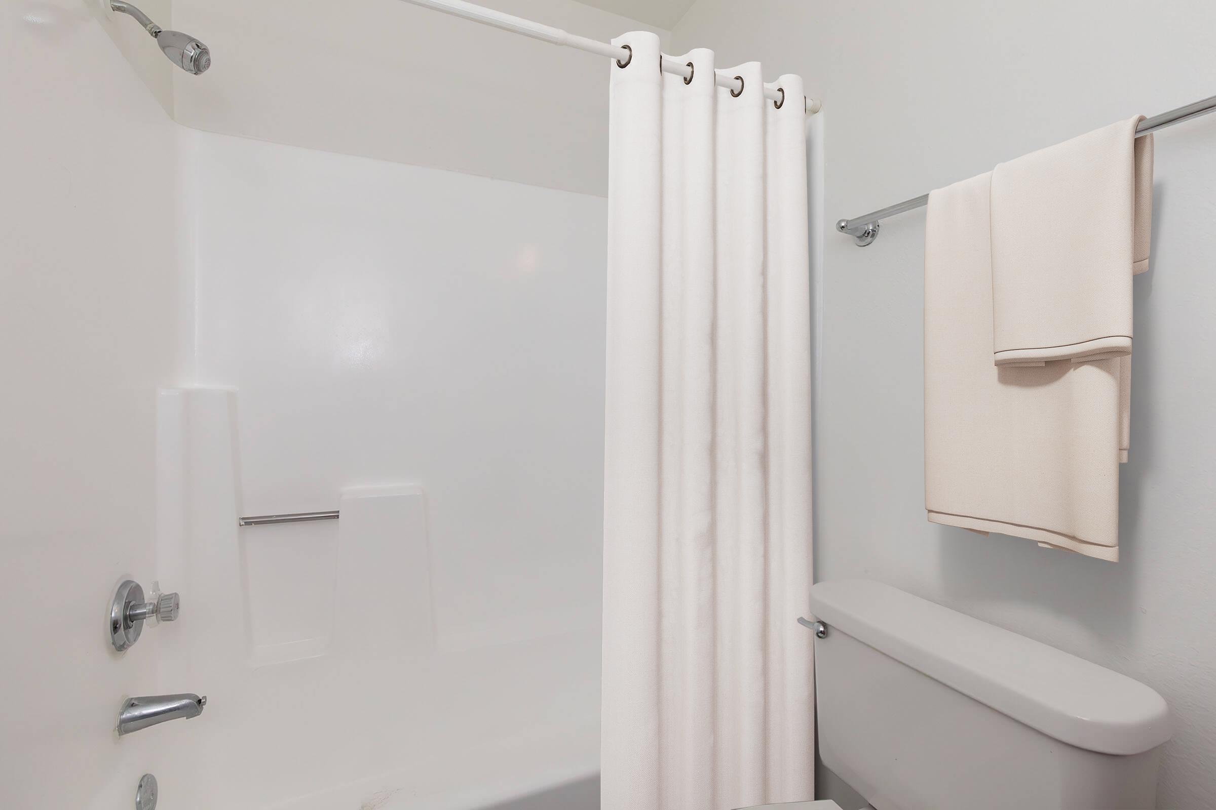 A clean and minimalistic bathroom featuring a bathtub with a white shower curtain, a towel rack with two neatly hung cream-colored towels, and a toilet. The walls are light-colored, creating a bright and fresh atmosphere.