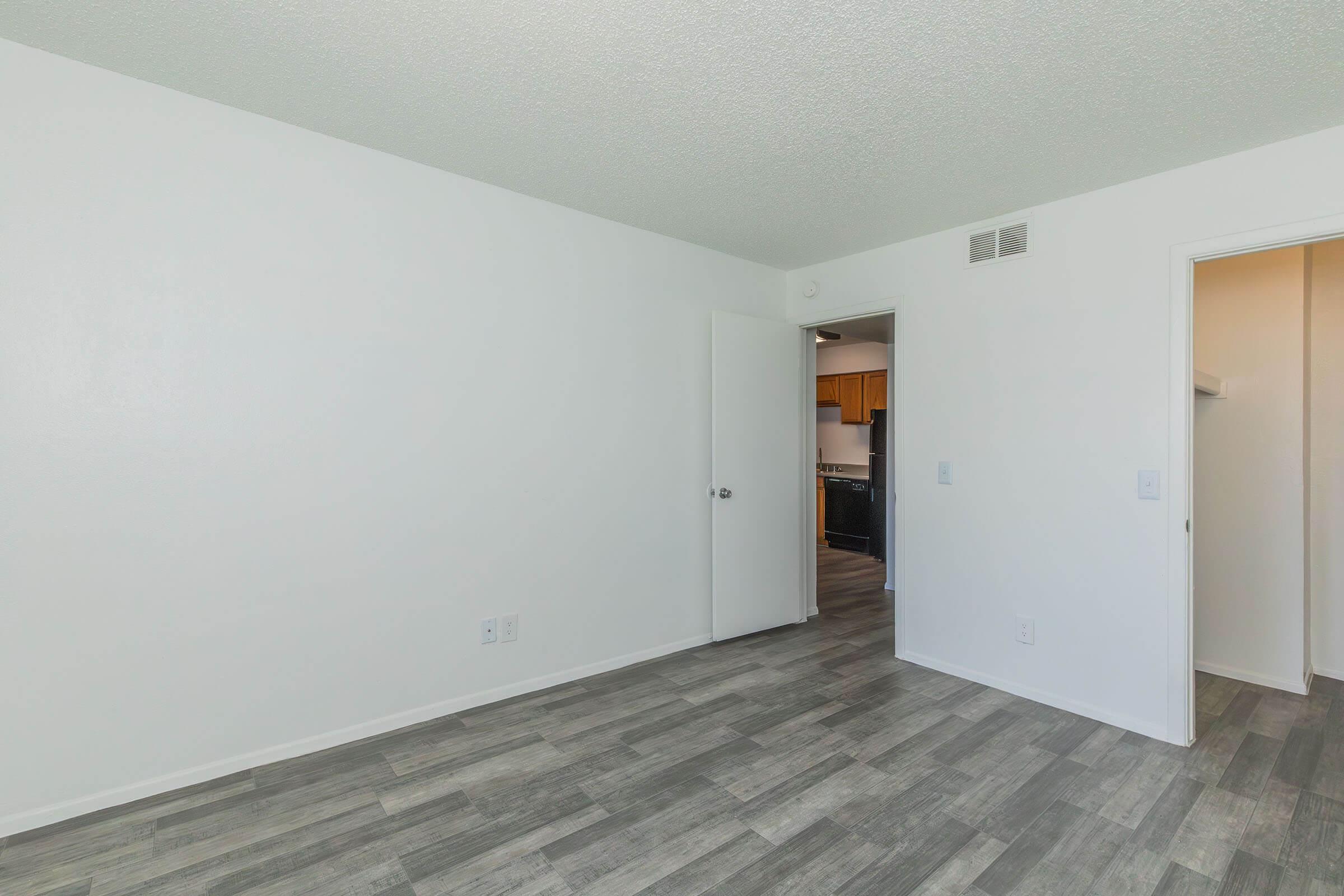 A clean, empty room with white walls and a textured ceiling. The floor features light gray tiles. A doorway on the left leads to a kitchen, which is partially visible. There is a closet space on the right with an adjacent door. The overall atmosphere is bright and spacious.