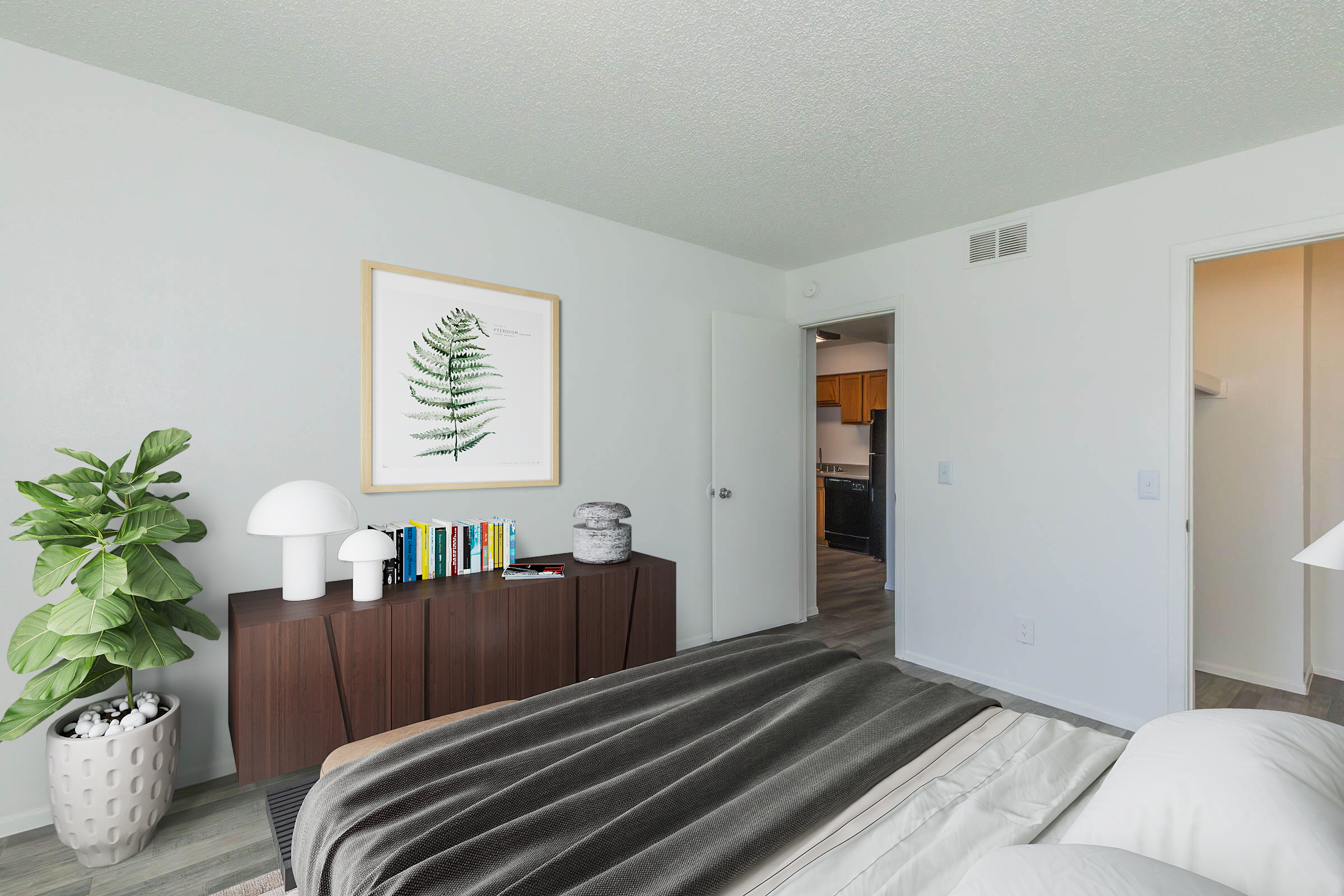 A modern bedroom featuring a neatly made bed with gray and cream bedding. A wooden dresser holds a stack of books, a decorative bowl, and a lamp, with a large framed fern print on the wall. A potted plant adds greenery to the space, and a doorway leads to another room in the background. The walls are painted white, enhancing the bright ambiance.