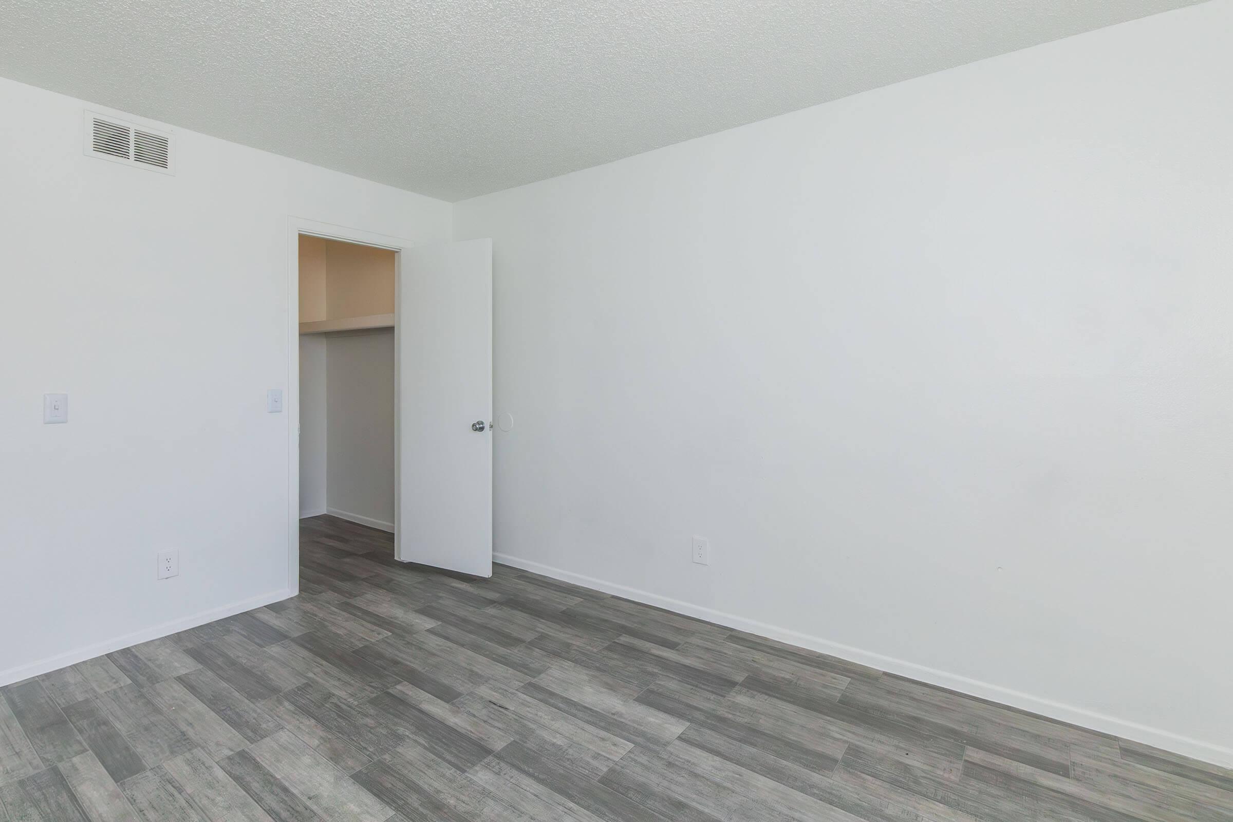 Empty room with light-colored walls and a wooden floor. A white door leads to a closet, and there's a door frame visible. The overall aesthetic is clean and minimalistic, with plenty of natural light. There are no furnishings or decorations present.