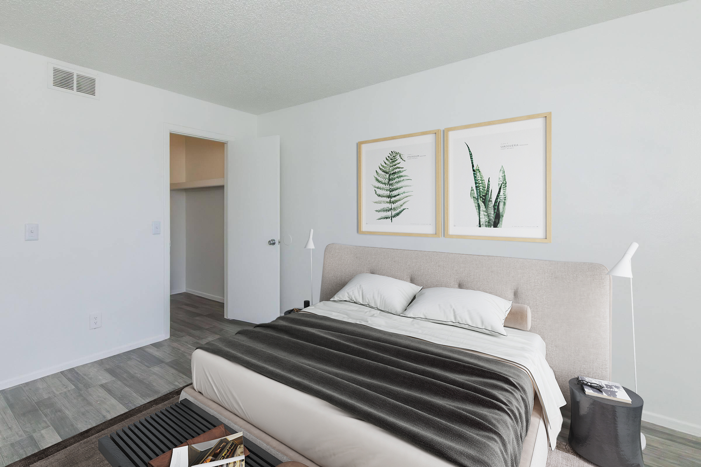 A modern bedroom featuring a large bed with a beige upholstered headboard, layered with a gray blanket. On the wall, there are two framed botanical prints. The room has light-colored walls and light wood flooring, with a small side table near the bed and a doorway visible leading to an adjacent room.