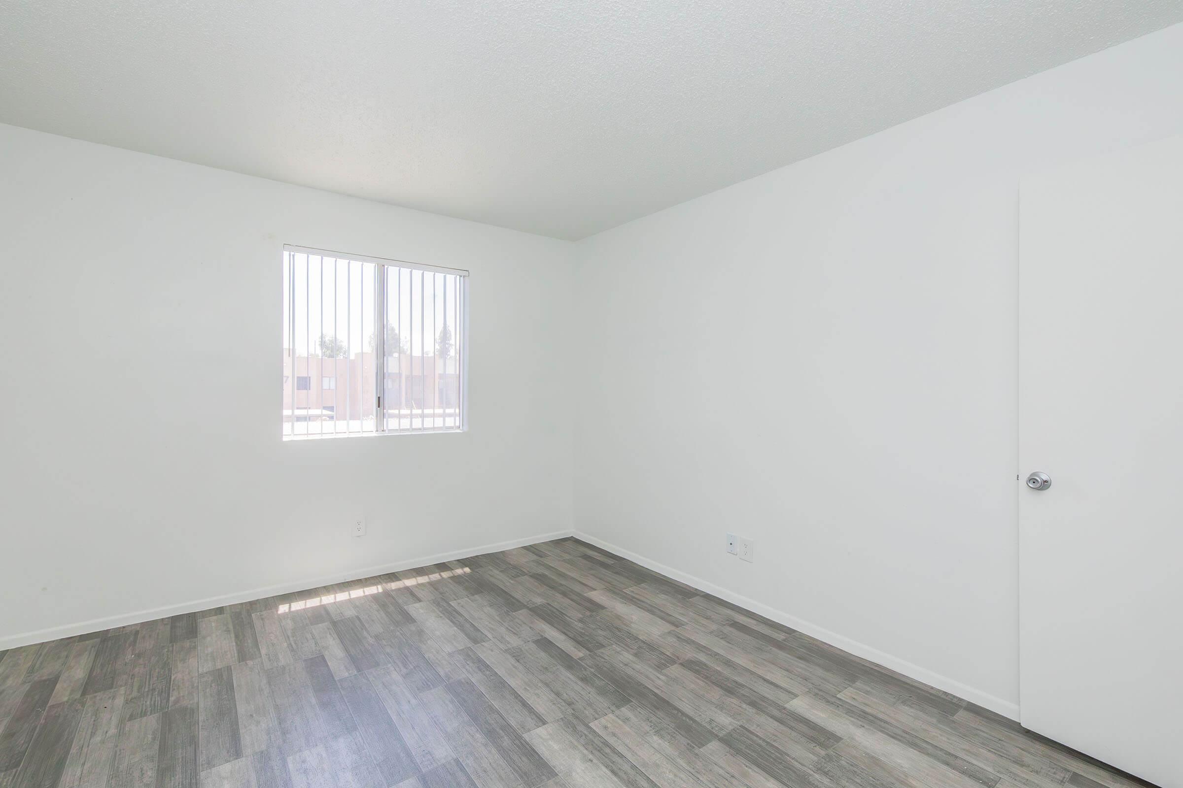 Empty room with white walls and a window featuring vertical blinds. The floor is covered in gray and brown laminate, and there is a door on the right side. The room has a bright and spacious feel, with natural light coming through the window.
