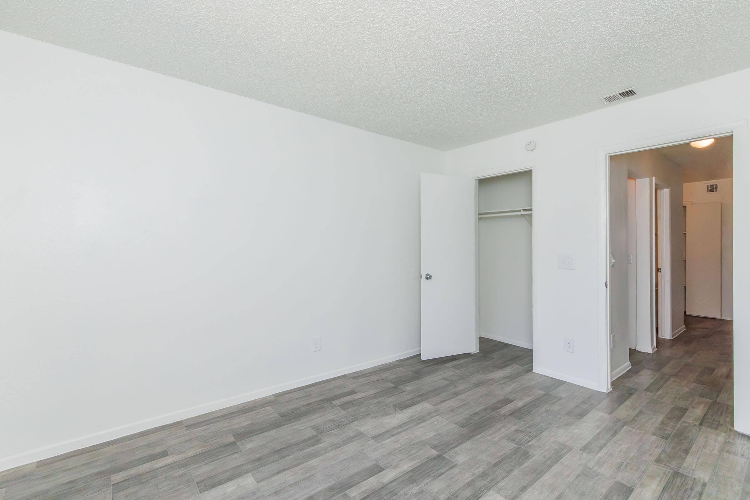 A bright, empty room with white walls and a textured ceiling. The floor features gray tile. There is an open closet with a sliding door on the left and a doorway leading to another area on the right. Natural light illuminates the space, creating a clean and minimalistic atmosphere.
