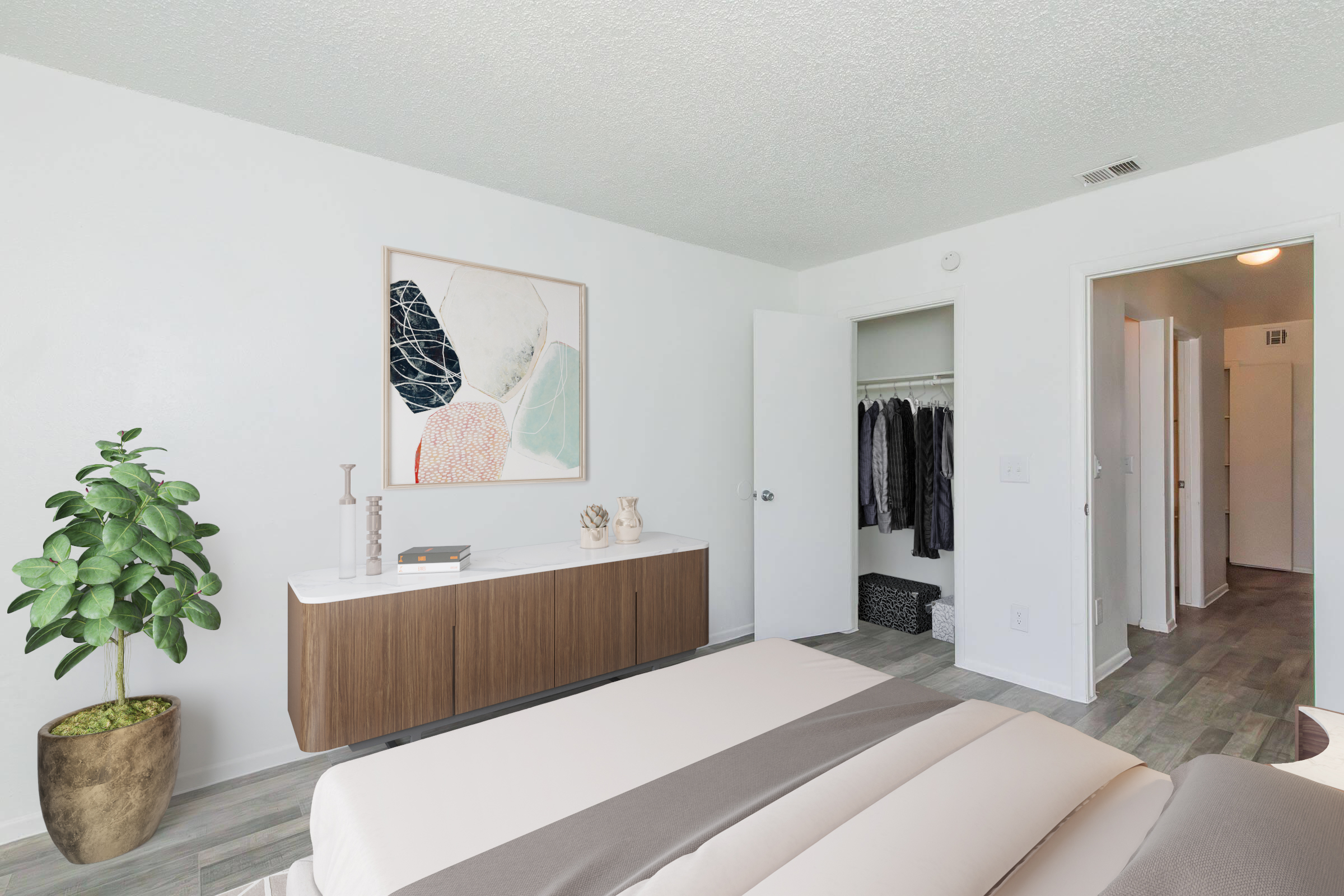 A modern, minimalist bedroom featuring a bed with beige and gray linens, a wooden side table, and a decorative plant. A large abstract painting is mounted on the wall. The room has a closet with hanging clothes visible through an open door and light-colored walls enhancing the airy atmosphere.