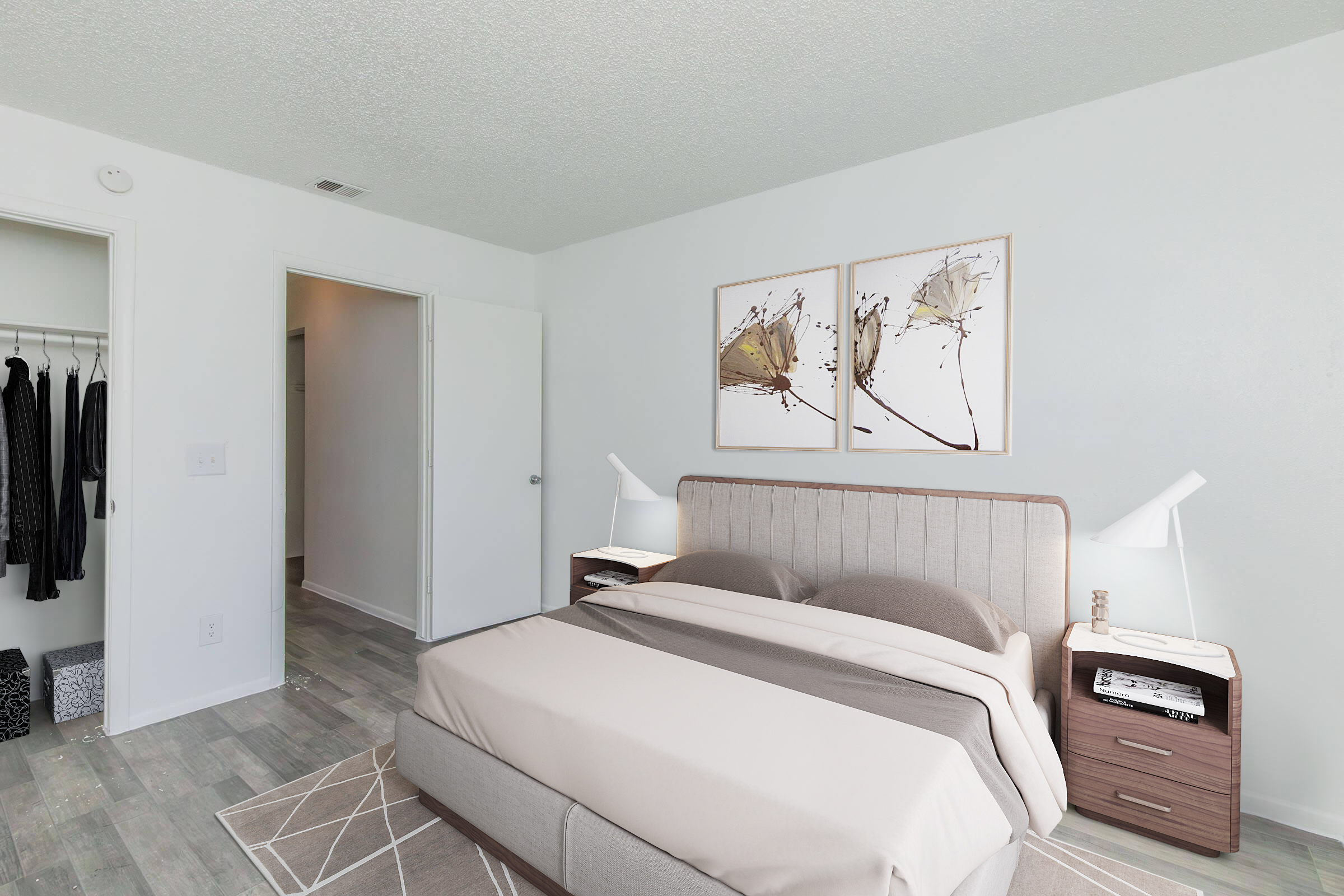 A modern bedroom featuring a beige bed with gray bedding, two wall art pieces above the headboard, and bedside tables with lamps. A light-colored closet is visible on the left, and a doorway leads to another room. The floor is covered with light wood planks, and a geometric area rug is placed beneath the bed.