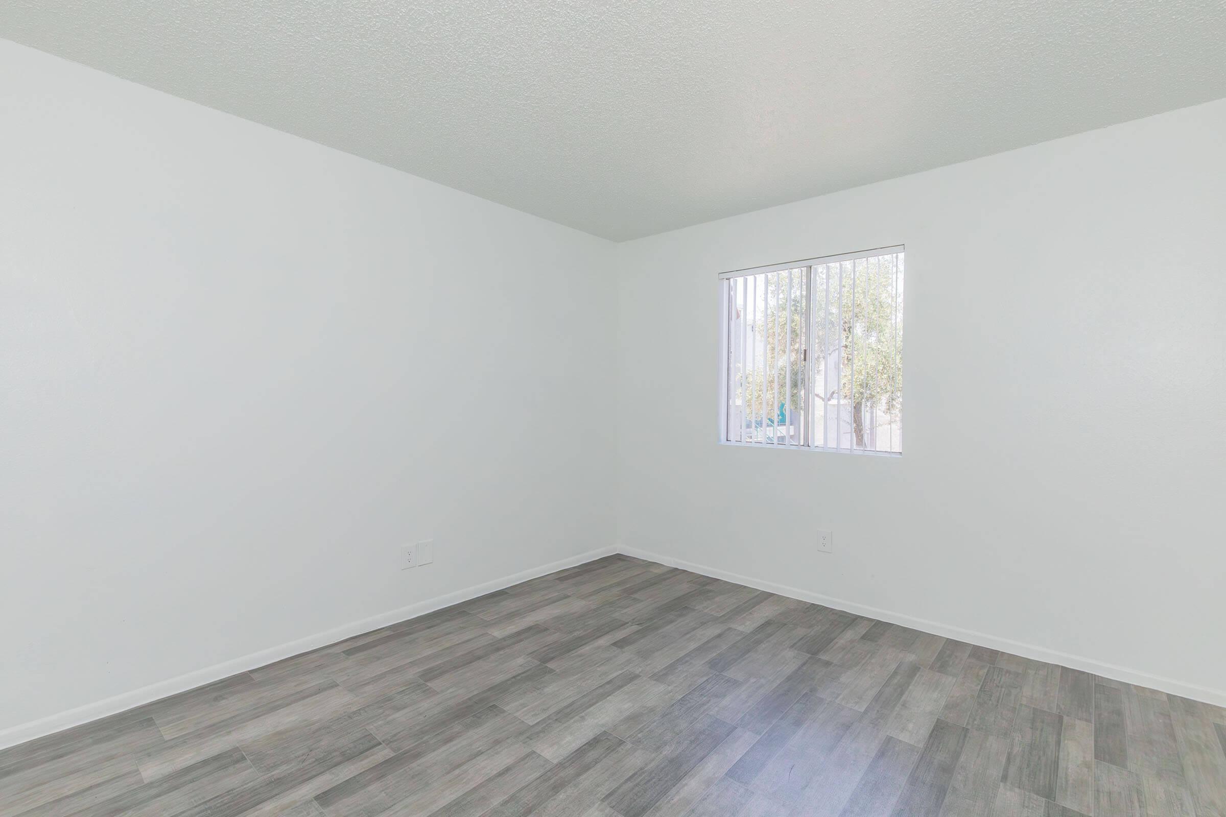 Empty room with white walls and a light gray laminate floor. A small window with vertical blinds allows natural light to enter. The space is unfurnished and clean, creating an open and minimalistic atmosphere.
