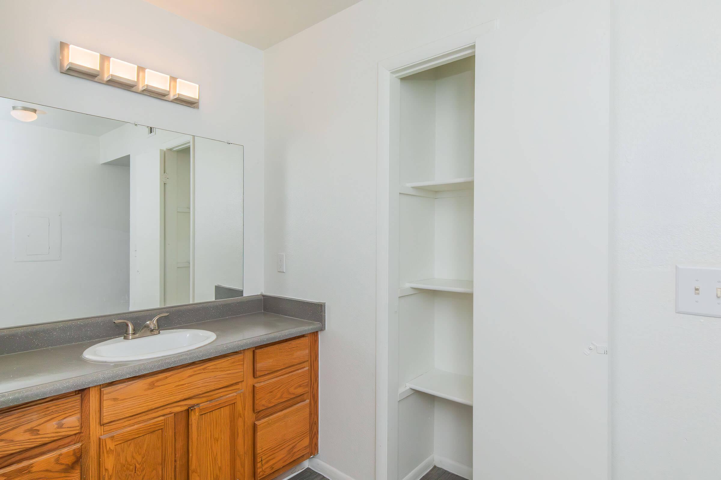 A bright bathroom featuring an oak vanity with a sink, a large mirror above the sink, and a wall-mounted light fixture. To the right, there is a closet with three shelves, and the walls are painted white, creating a clean and spacious feel. The floor is tiled in a light shade.