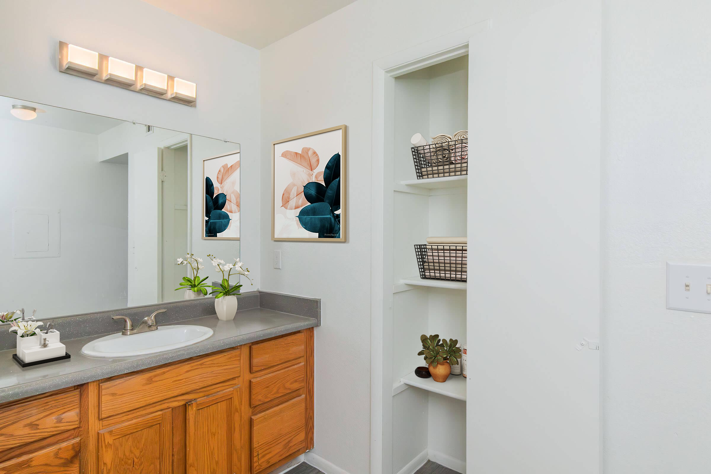 A modern bathroom featuring a gray countertop with a sink, wooden cabinetry, and a wall-mounted mirror. There is a decorative wall art piece beside the mirror, and a small shelf unit with woven baskets. The space is well-lit and has a clean, minimalist aesthetic.