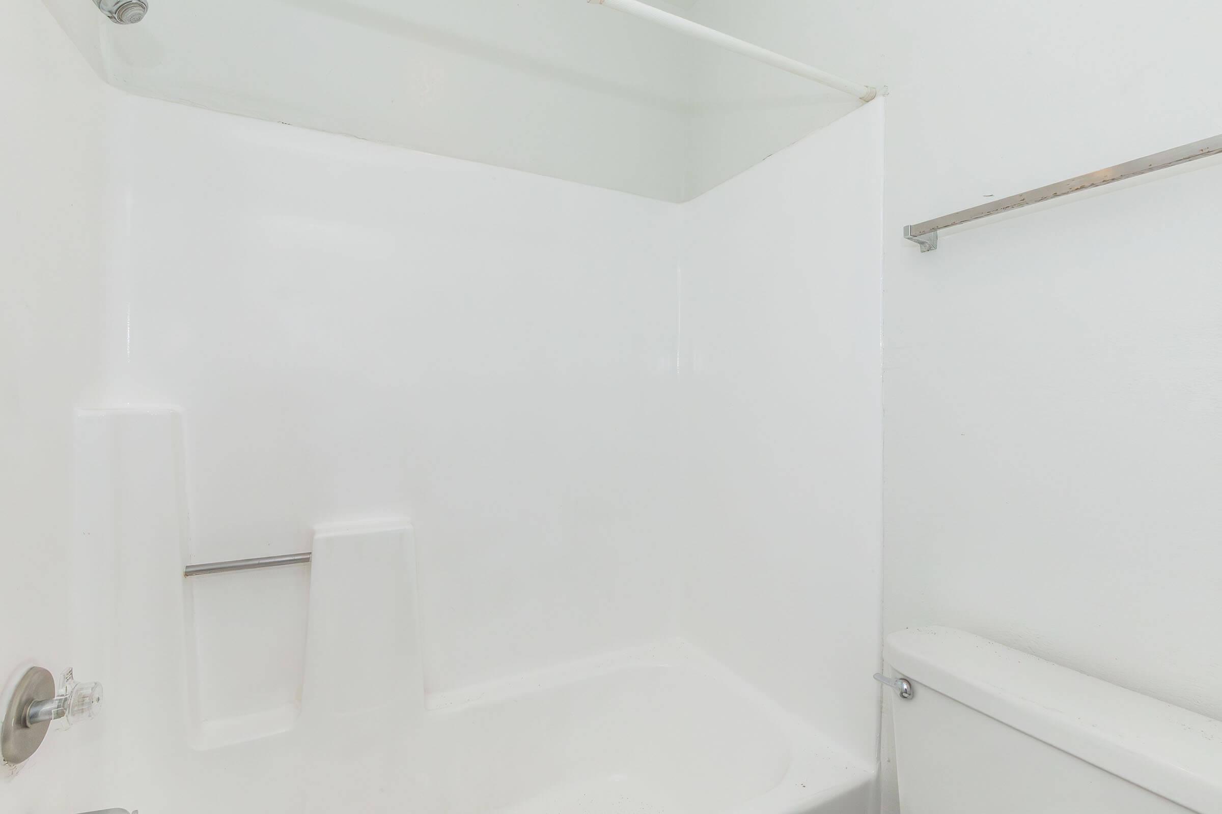 A clean, minimalist bathroom featuring a white tub and shower enclosure, a toilet, and a bare towel rack. The walls are painted white, contributing to a bright and spacious feel.