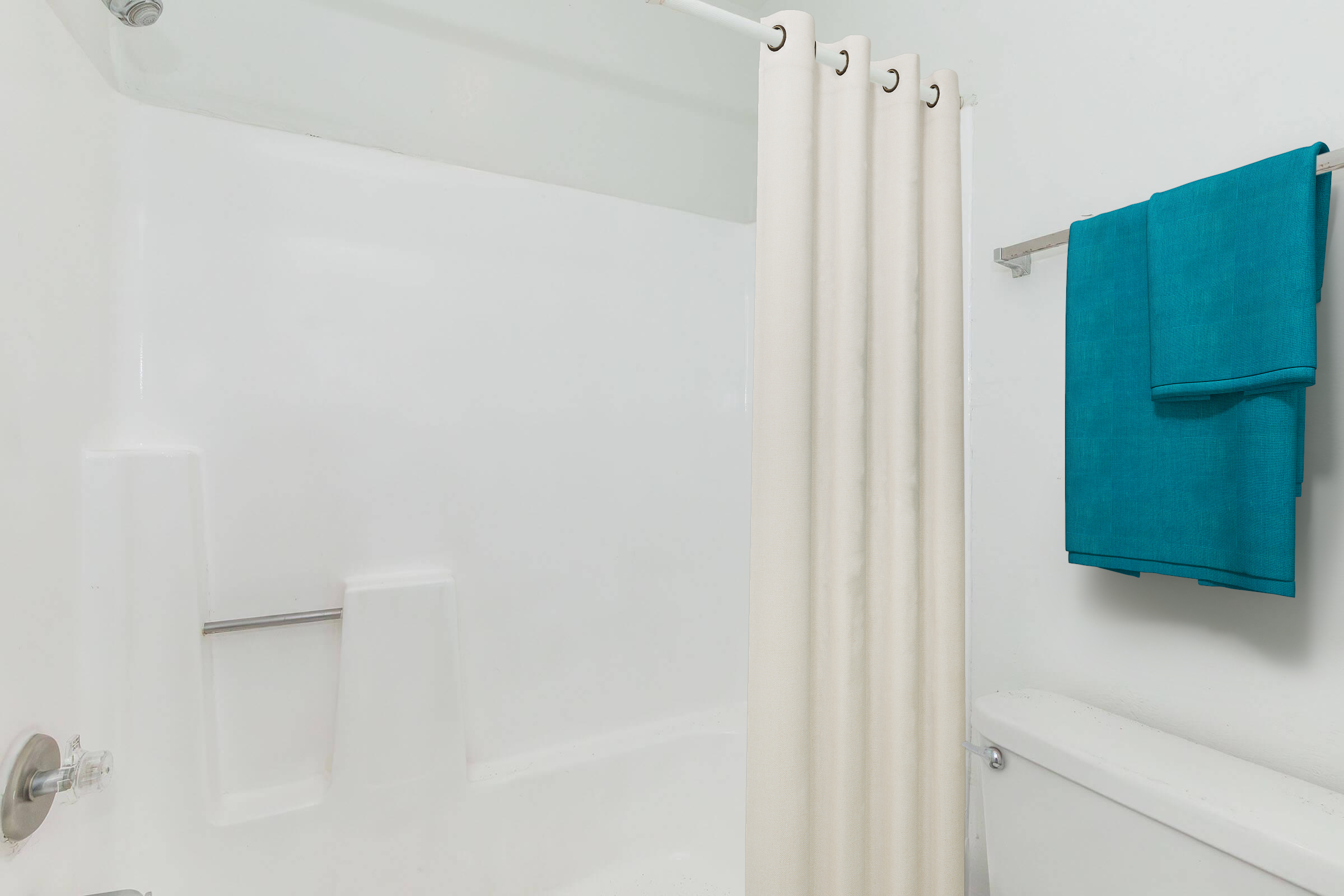 A clean and simple bathroom setting featuring a white bathtub and wall. There is a light-colored shower curtain hanging from a rod, and two vibrant turquoise towels neatly arranged on a metal rack near the toilet, adding a pop of color to the space. The overall aesthetic is bright and minimalist.