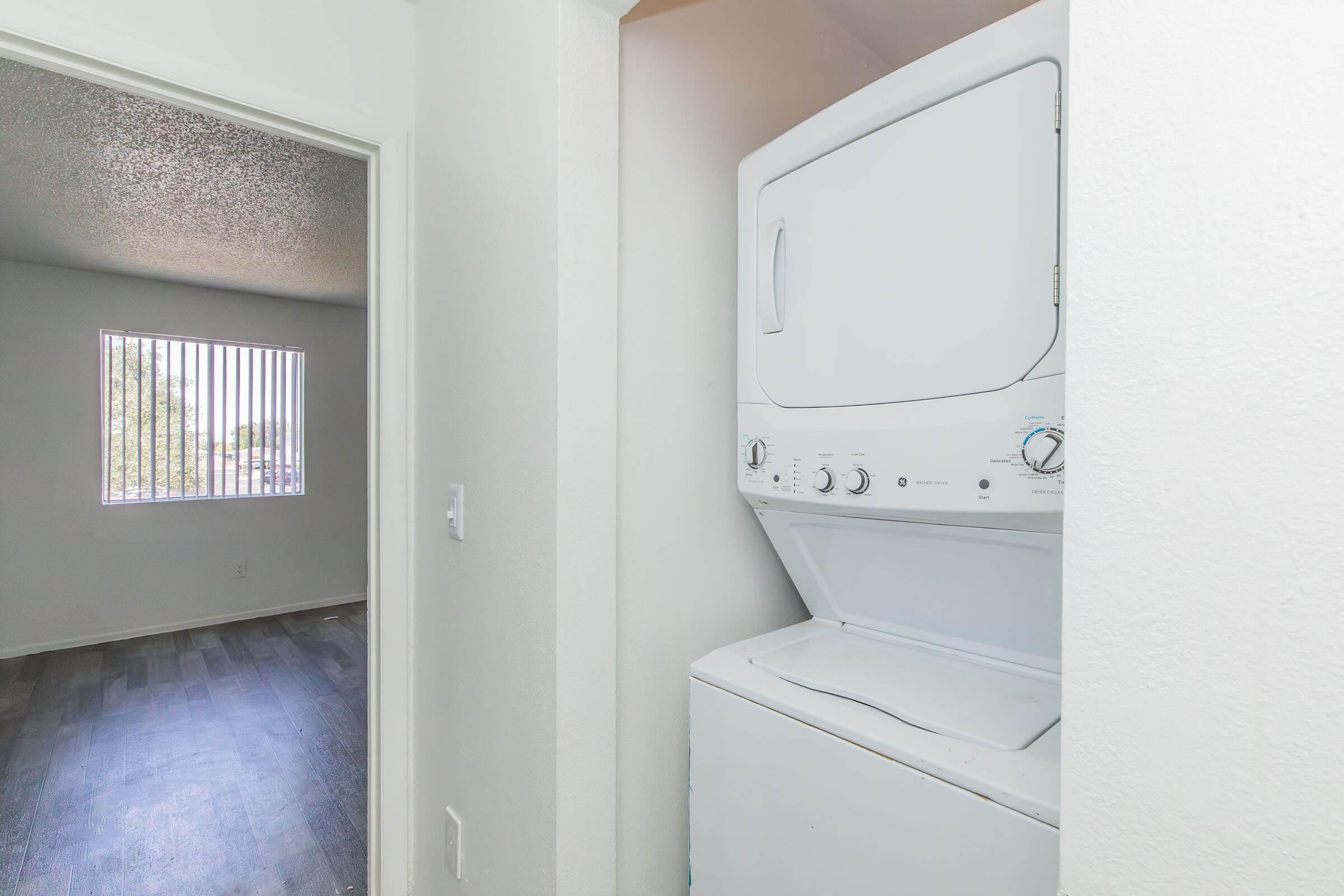 A stacked washing machine and dryer unit in a small laundry space, with a doorway leading to an adjacent room featuring a window with blinds. The walls are painted in a light color, and the flooring appears to be hardwood or laminate.