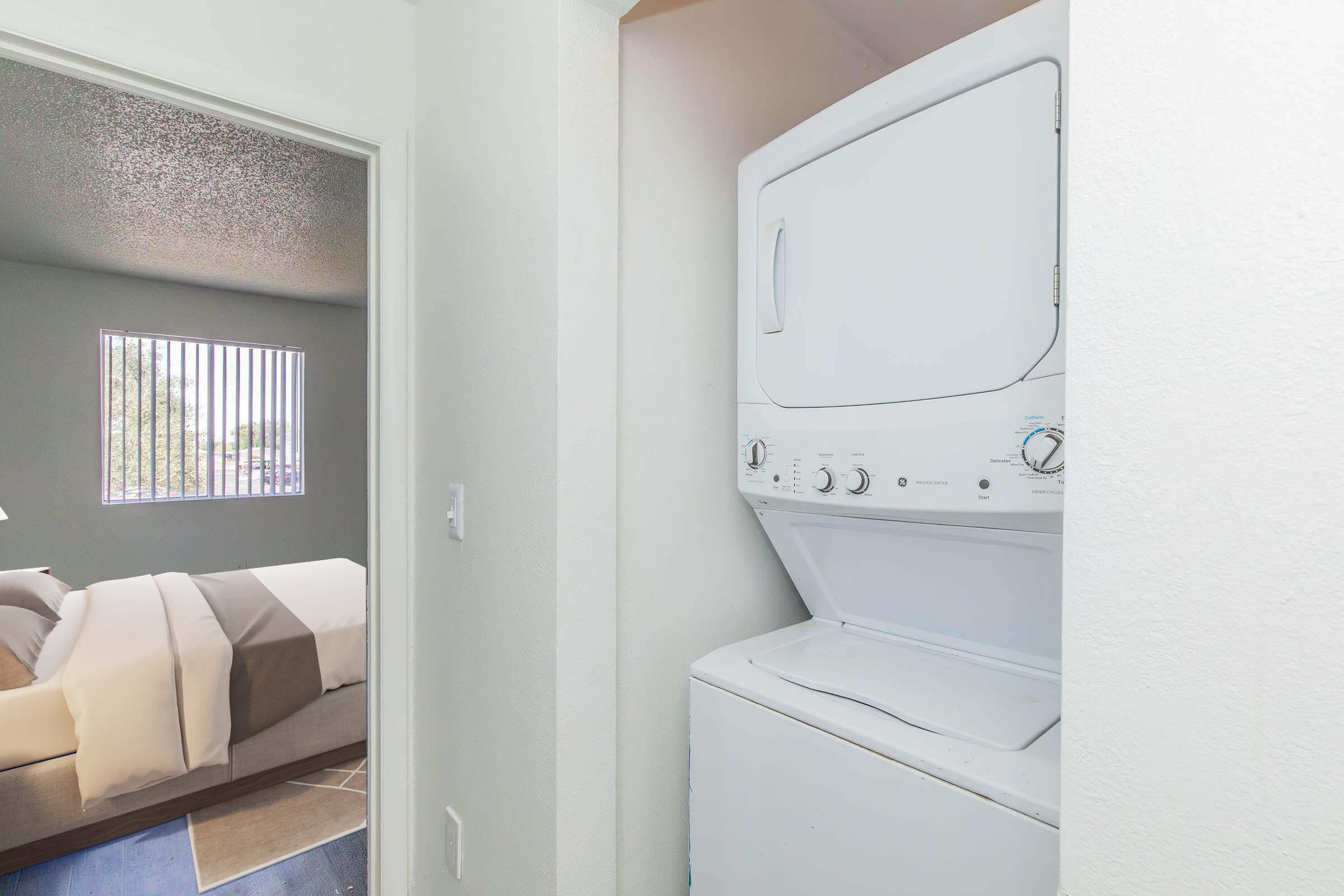 A laundry area featuring a stacked washer and dryer, positioned in a small space near a doorway. In the background, a well-lit bedroom with a window showcasing vertical blinds and a bed dressed in neutral-toned bedding is visible.