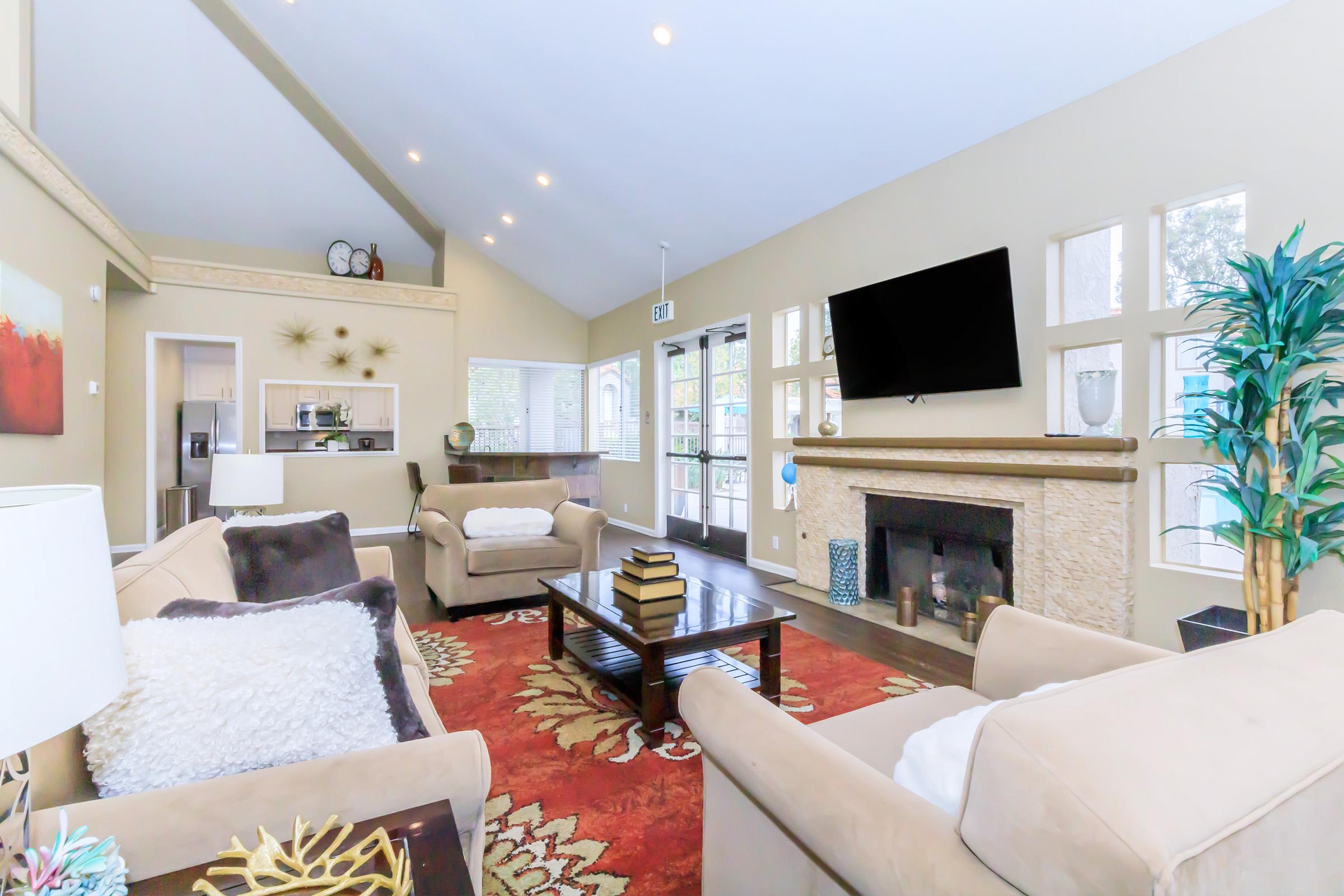 A bright and spacious living room featuring two beige sofas with decorative pillows, a central coffee table with stacked books, and a large area rug. There are windows allowing natural light, a fireplace, and a television mounted on the wall, creating a cozy and inviting atmosphere.