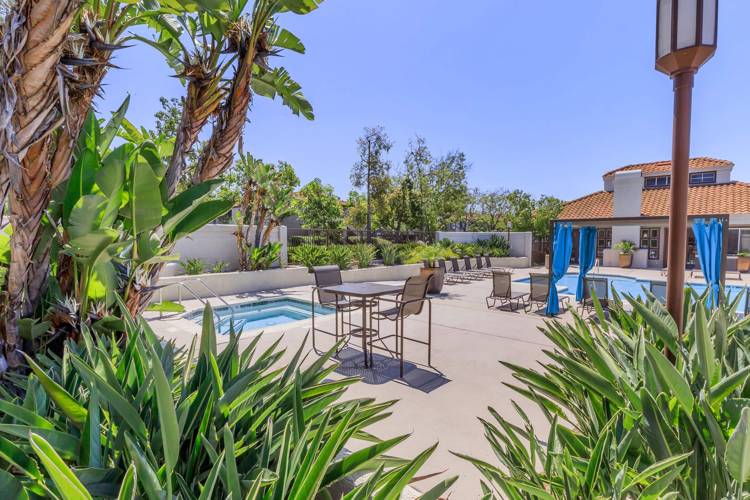 A sunny outdoor pool area featuring a swimming pool, lounge chairs, and a hot tub. Surrounded by tropical plants and palm trees, with blue skies overhead. The space includes tables and chairs for seating, creating a relaxing atmosphere. There are also large blue curtains by the poolside for added shade.
