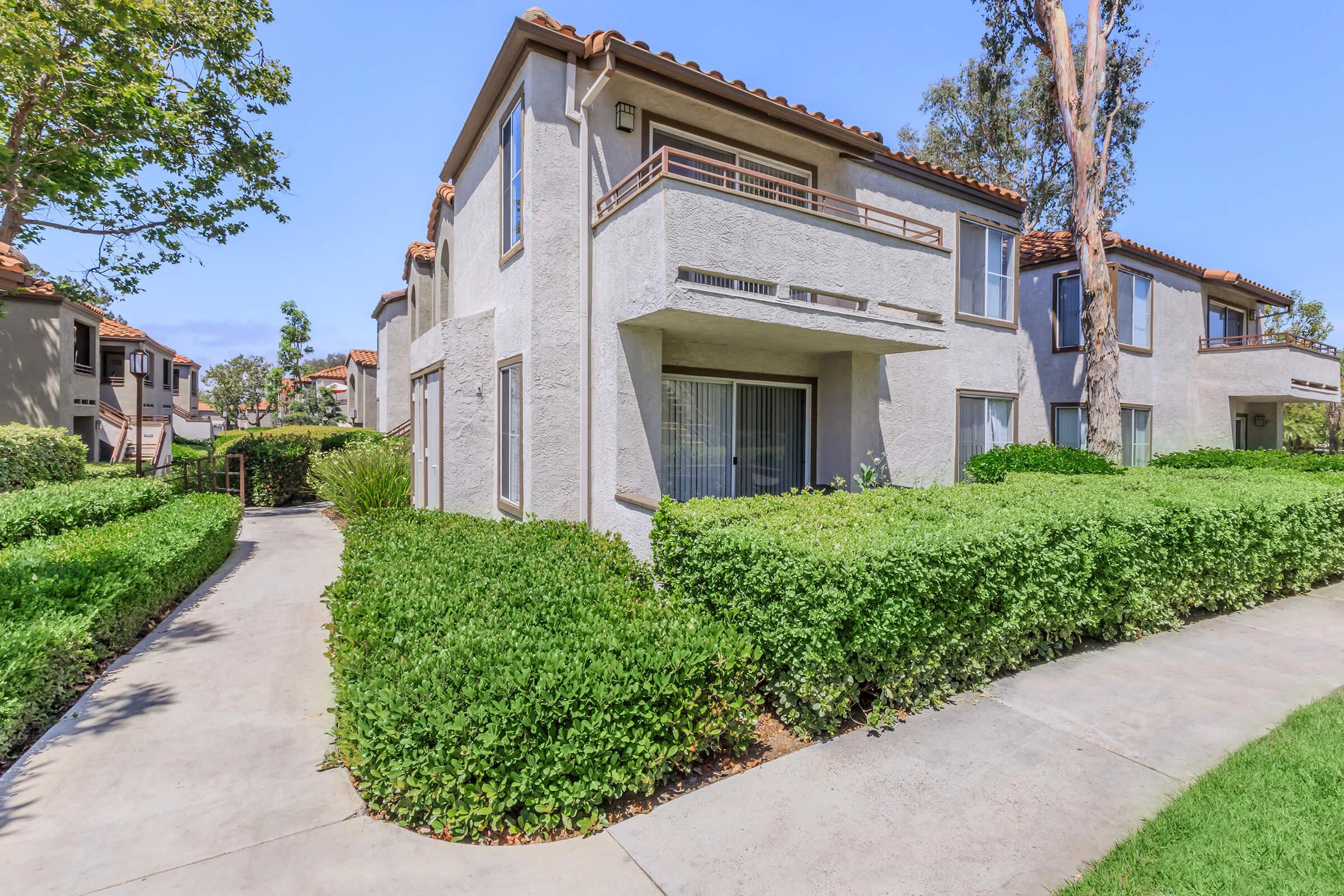 Two-story apartment building with a light gray exterior, situated among lush green landscaping. The pathway leads to the entrance, bordered by neatly trimmed hedges. Large windows provide a view of the interior, and trees are visible in the surrounding area, enhancing the inviting atmosphere.