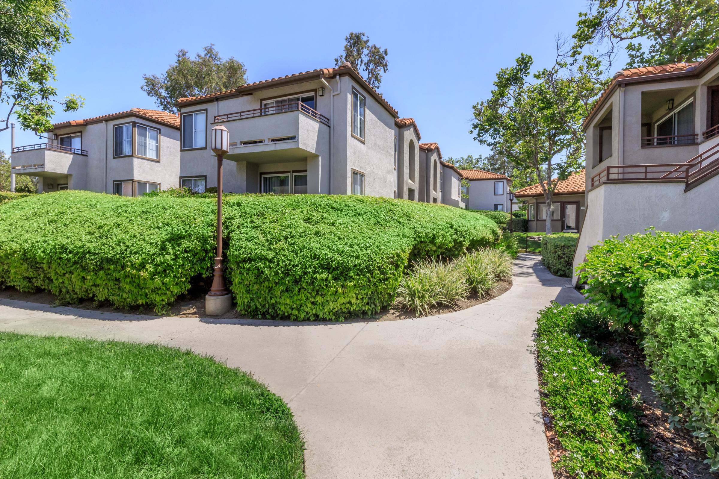 A view of a landscaped pathway winding through a residential area, featuring two-story apartment buildings surrounded by lush green shrubbery and trees. The buildings have a Mediterranean architectural style, with terracotta roofs and balconies. The scene is bright and sunny, indicating a pleasant environment.
