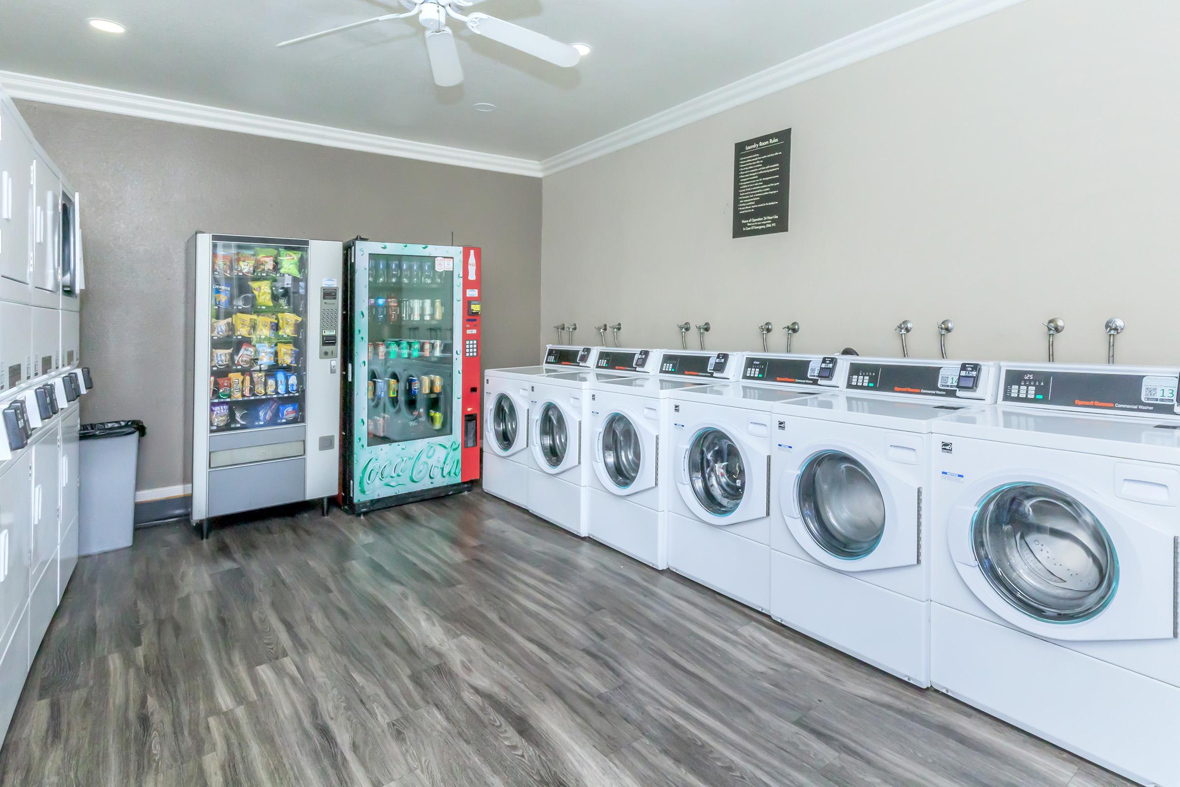 A bright and clean laundry room featuring several white washing machines and dryers lined up against the wall. There is a vending machine with snacks and drinks in the corner, and a large window allowing natural light to enter the space. The flooring is a modern wood laminate.