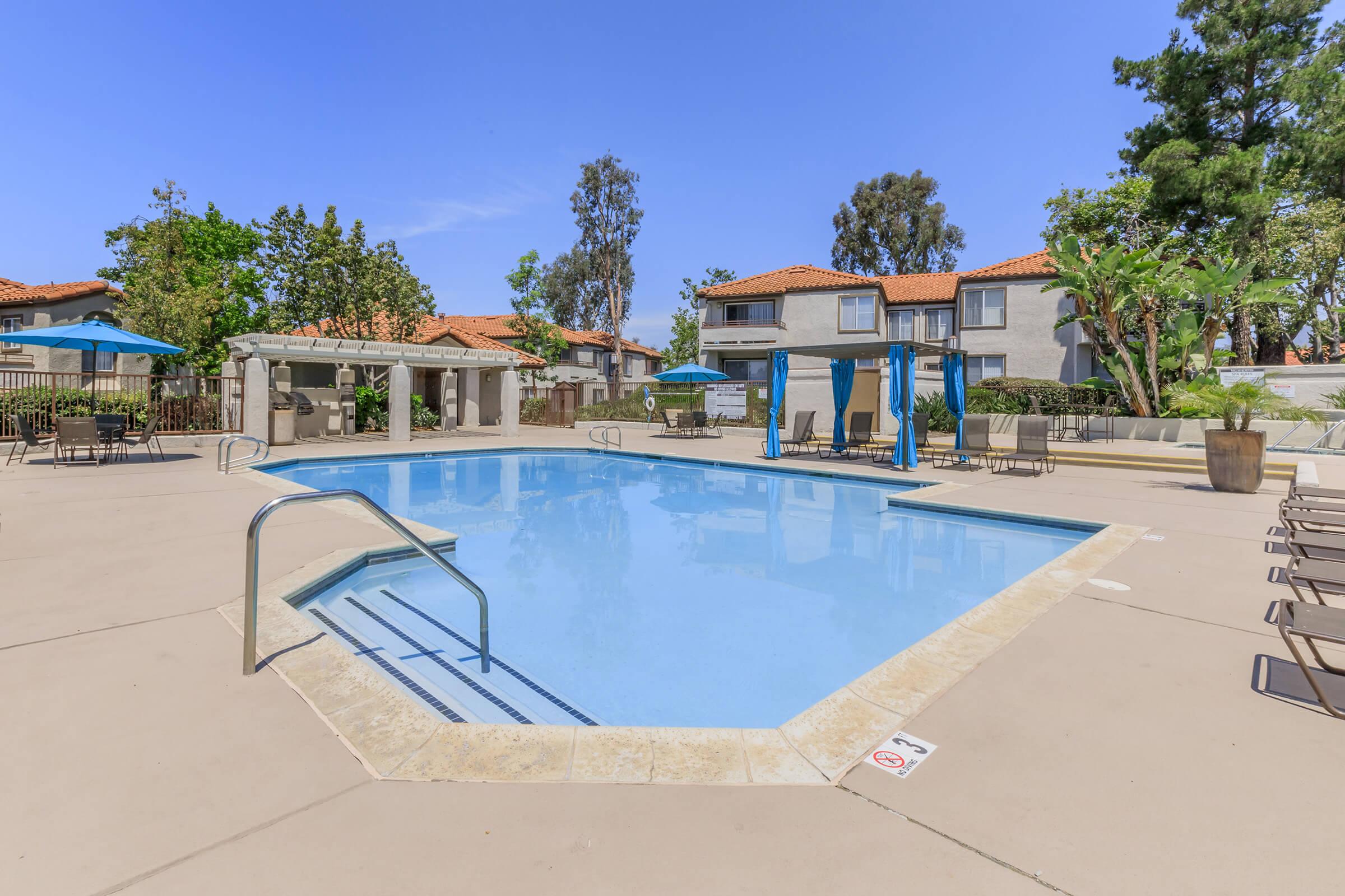A sunny outdoor pool area featuring a clear blue swimming pool surrounded by lounge chairs and umbrellas. Nearby are landscaped trees and residential buildings with red-tiled roofs, creating a relaxing atmosphere for leisure and recreation.