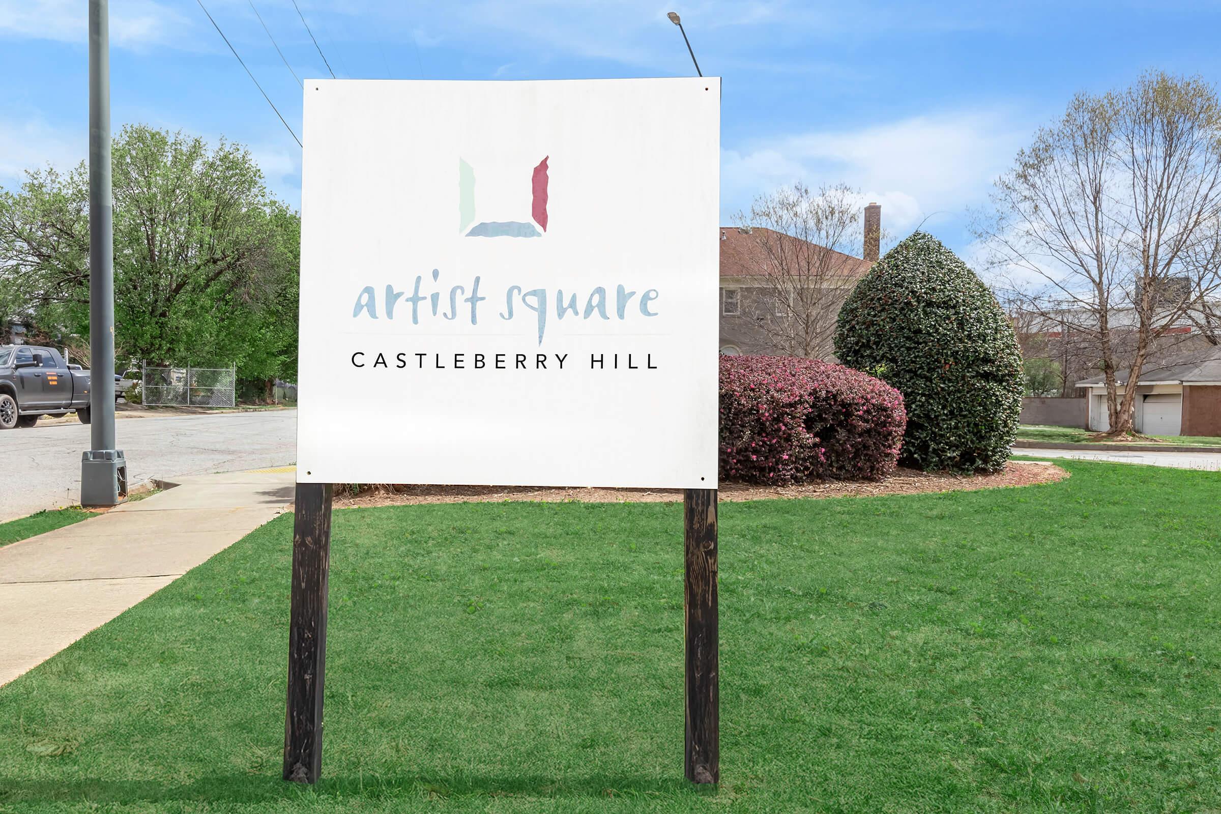 Sign for Artist Square in Castleberry Hill, featuring a simple logo with colorful accents. The sign is placed on green grass with shrubs in the background, under a clear blue sky.