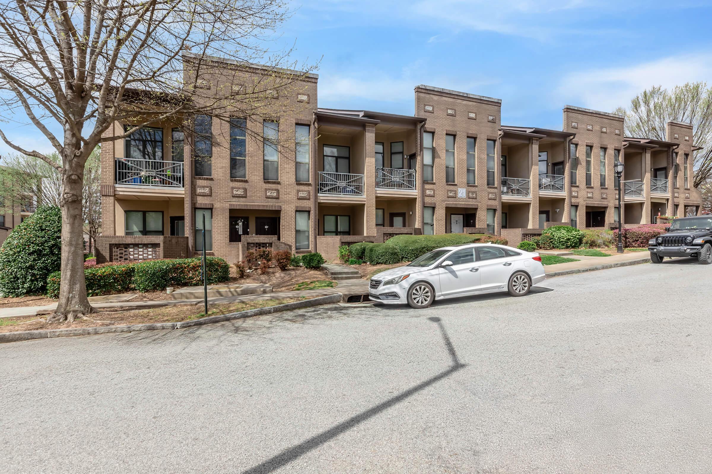 A row of modern, multi-unit townhouses with balconies, set along a tree-lined street. There are several parked cars, including a white sedan and a black SUV. Lush greenery and shrubs surround the buildings, creating a pleasant residential atmosphere.