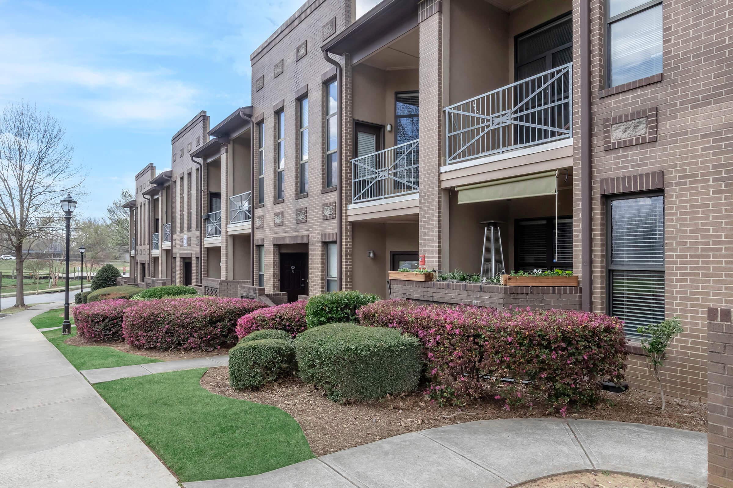 A row of modern brick apartments featuring balconies, surrounded by well-maintained shrubs and colorful flowering plants. A paved walkway leads through the landscaped area, with a tree visible in the background and clear blue skies above.